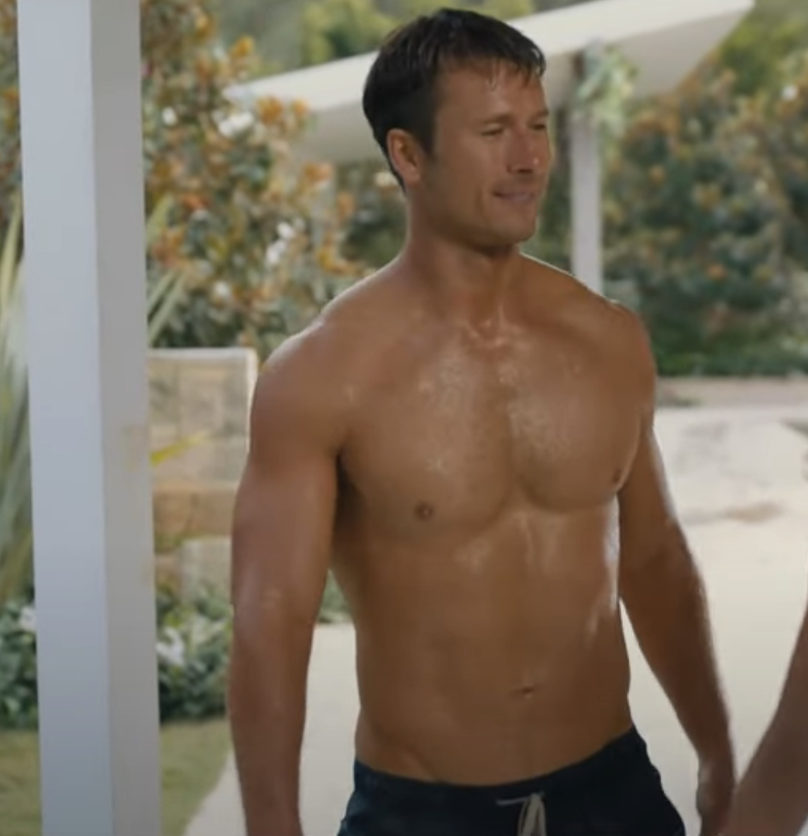 A shirtless man stands outdoors by a pool, smiling. He is wearing dark swimming trunks