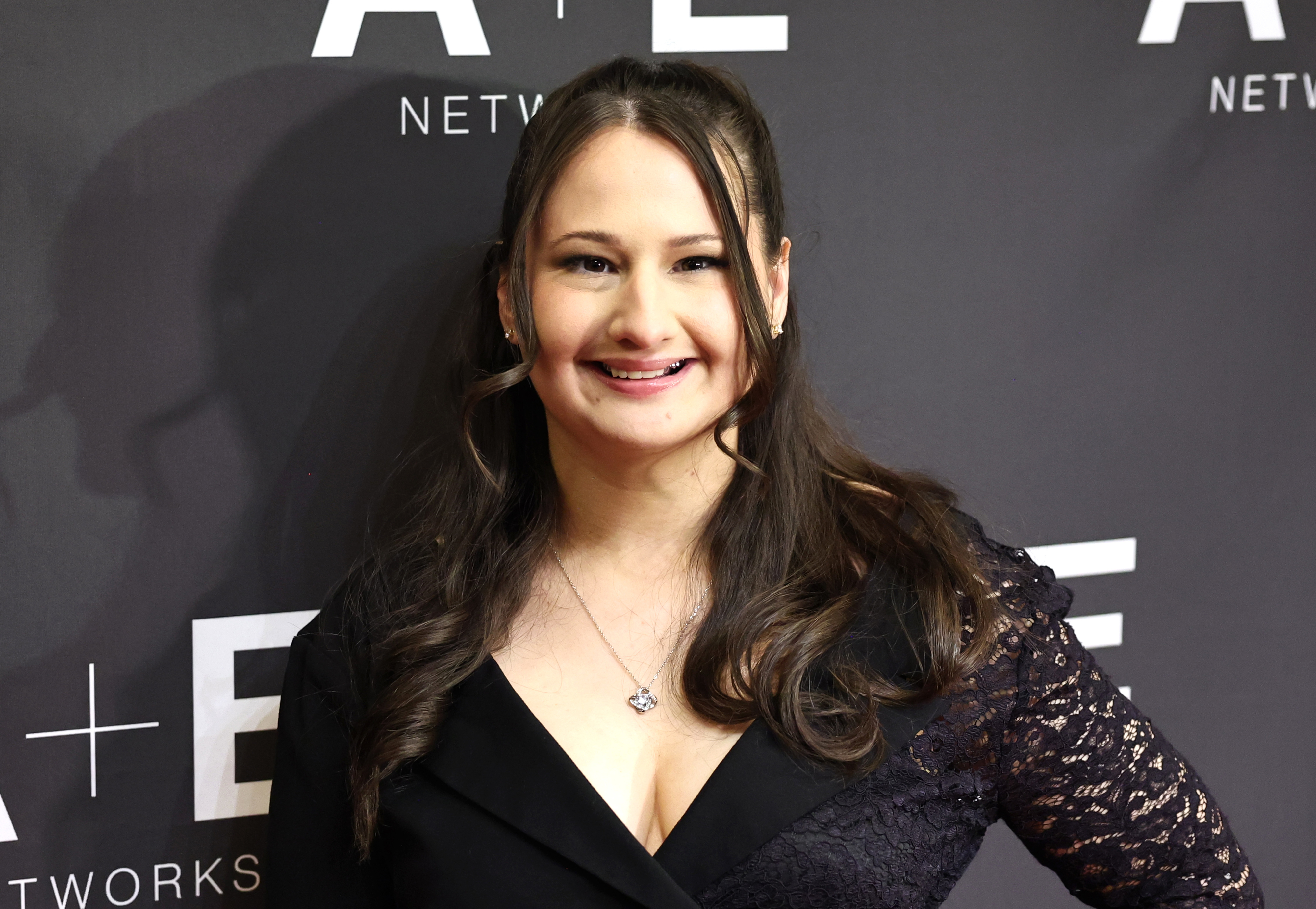 A woman smiles at the camera, wearing a detailed gown with lace sleeves at a red carpet event