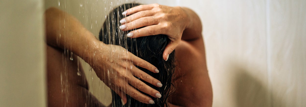 A person is seen from the back with their hands in their hair, standing under a shower stream