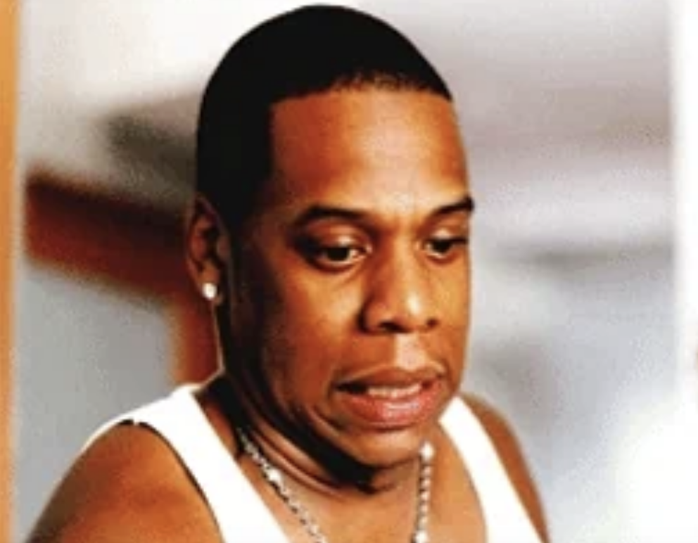 Jay-Z, wearing a sleeveless top and a necklace, appears pensive, looking down with a slightly furrowed brow in this candid image