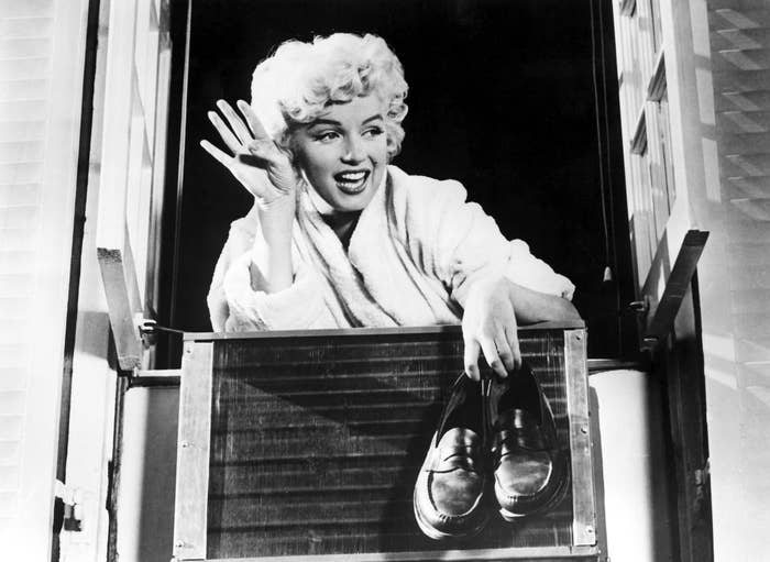 Marilyn Monroe waves while leaning out of a window, holding a pair of shoes, and wearing a robe