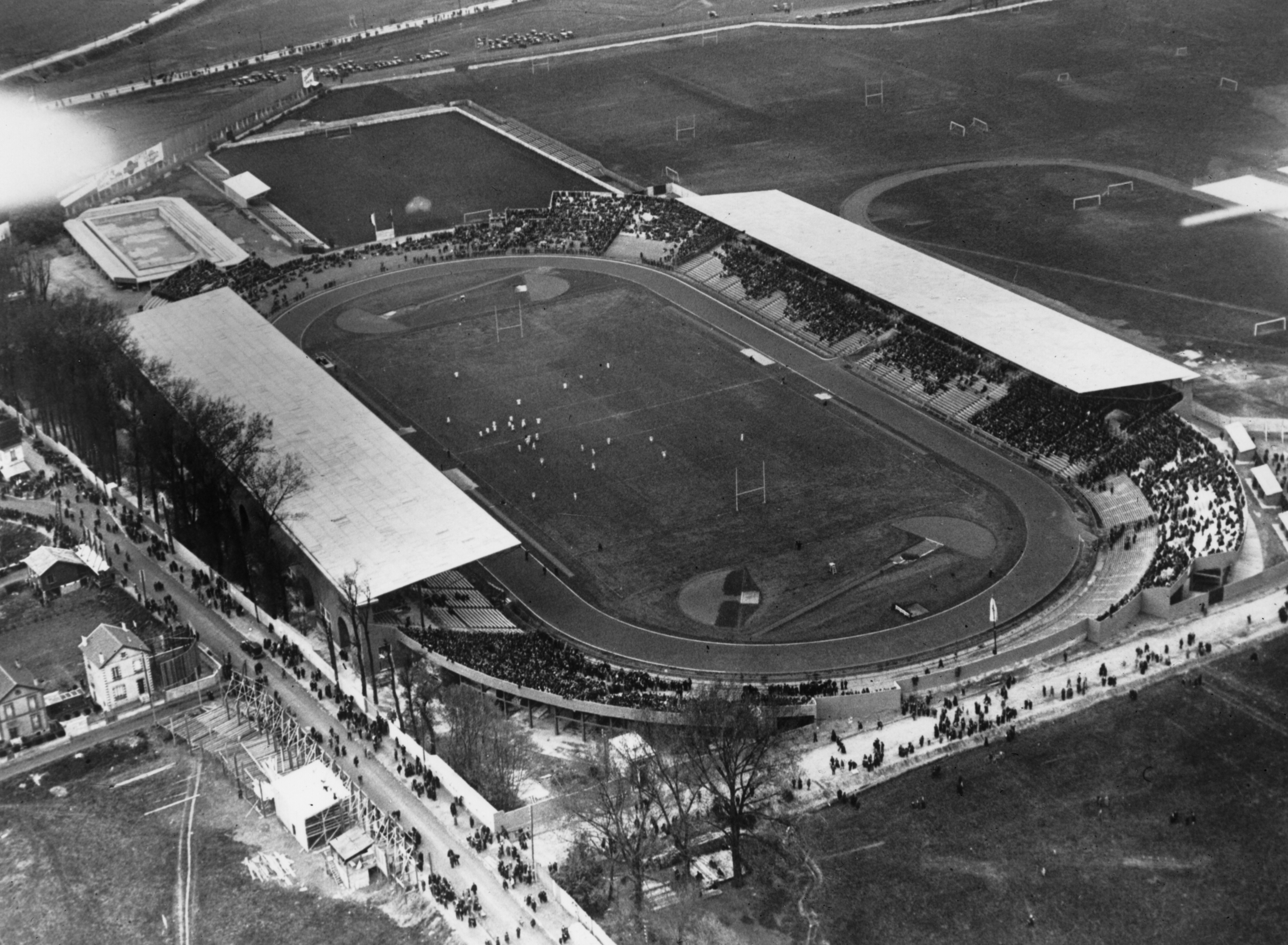 Aerial view of a large sports stadium with a track and field, spectators in the stands, and adjacent buildings and fields