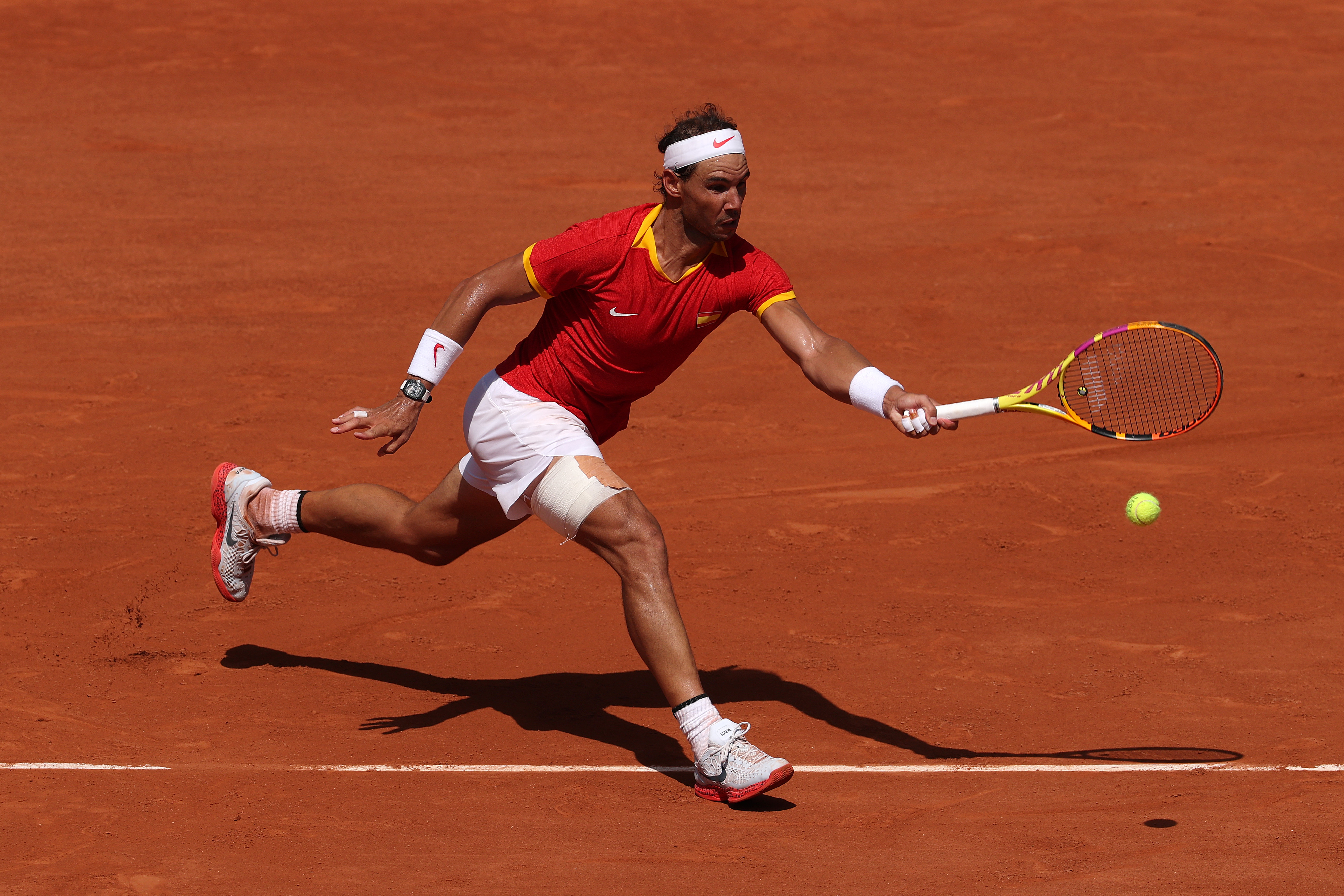 Rafael Nadal playing tennis on a clay court, wearing a red and yellow outfit, white headband, and white shoes, as he lunges to hit the ball