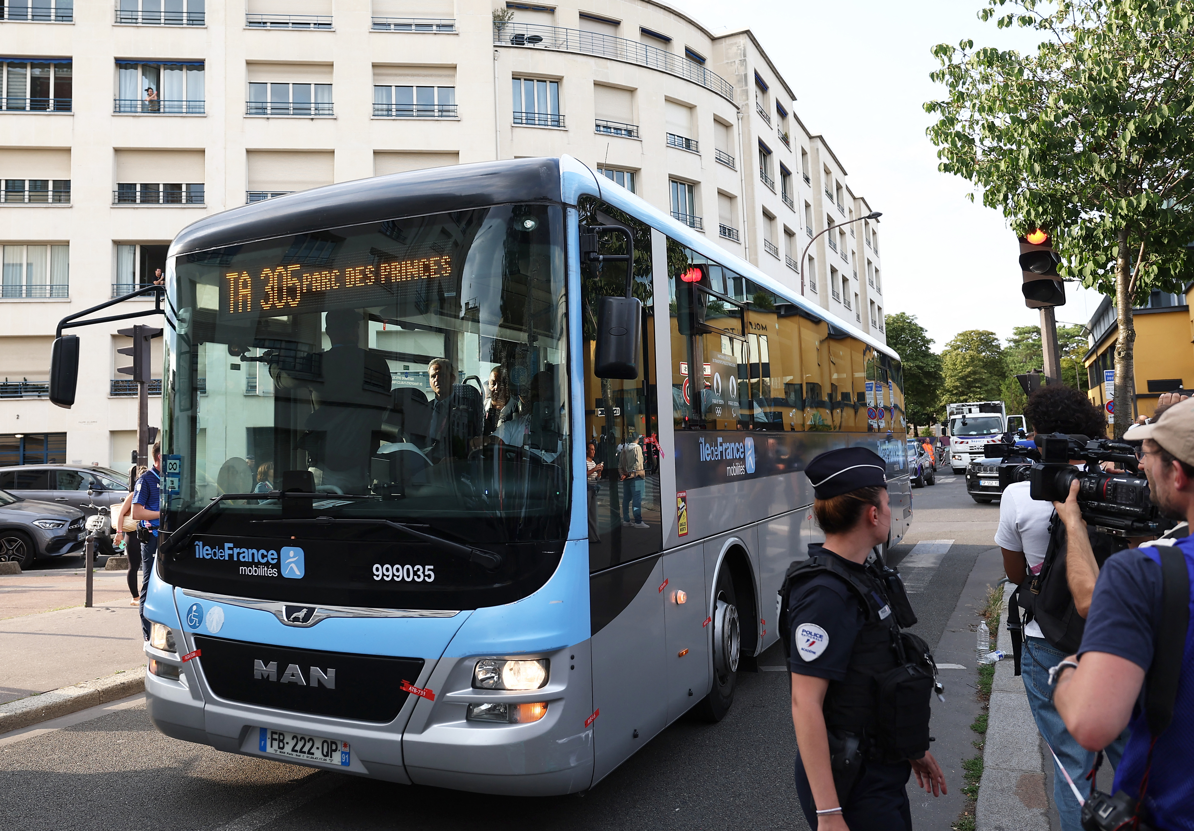 A football team bus arrives at a stadium, photographed by journalists and fans in an urban setting