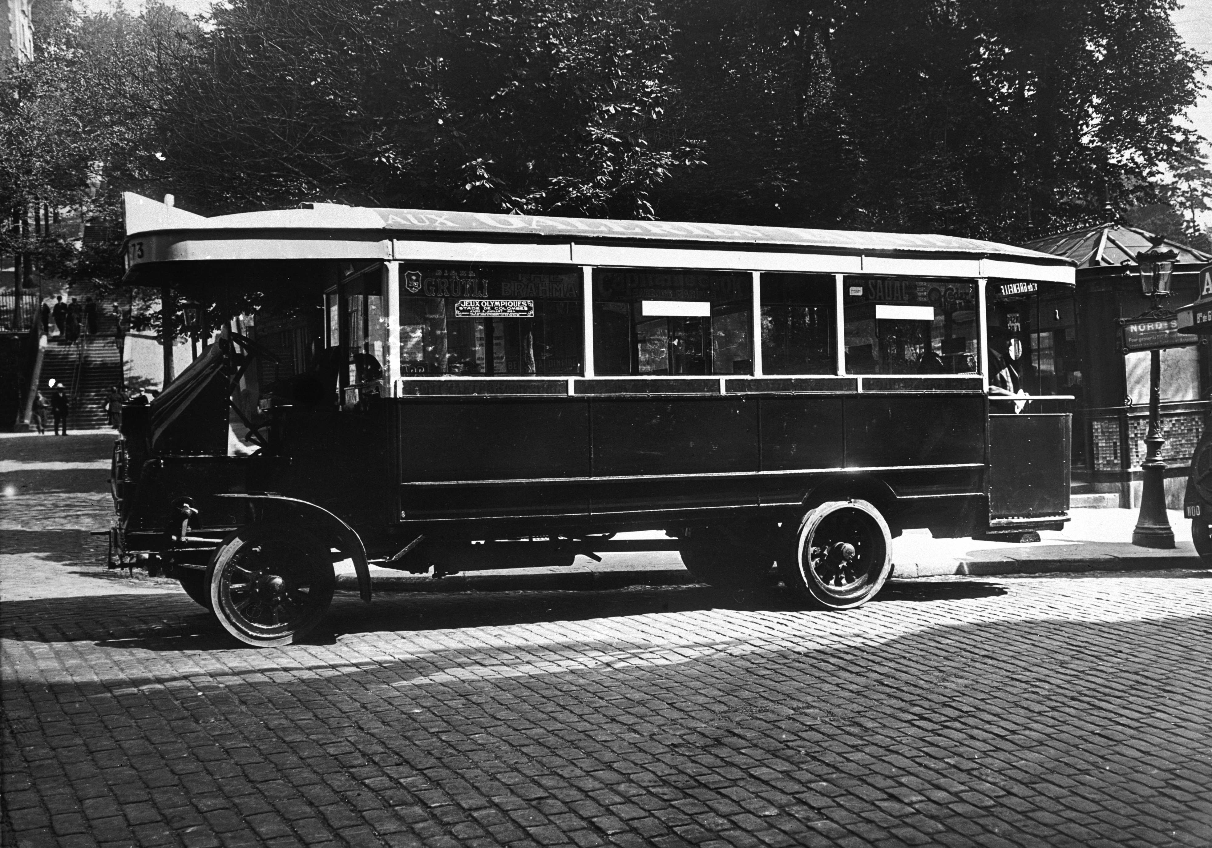 Black and white photo of an early 20th-century bus stopped at a cobblestone street, trees and buildings in the background. No text visible in the image