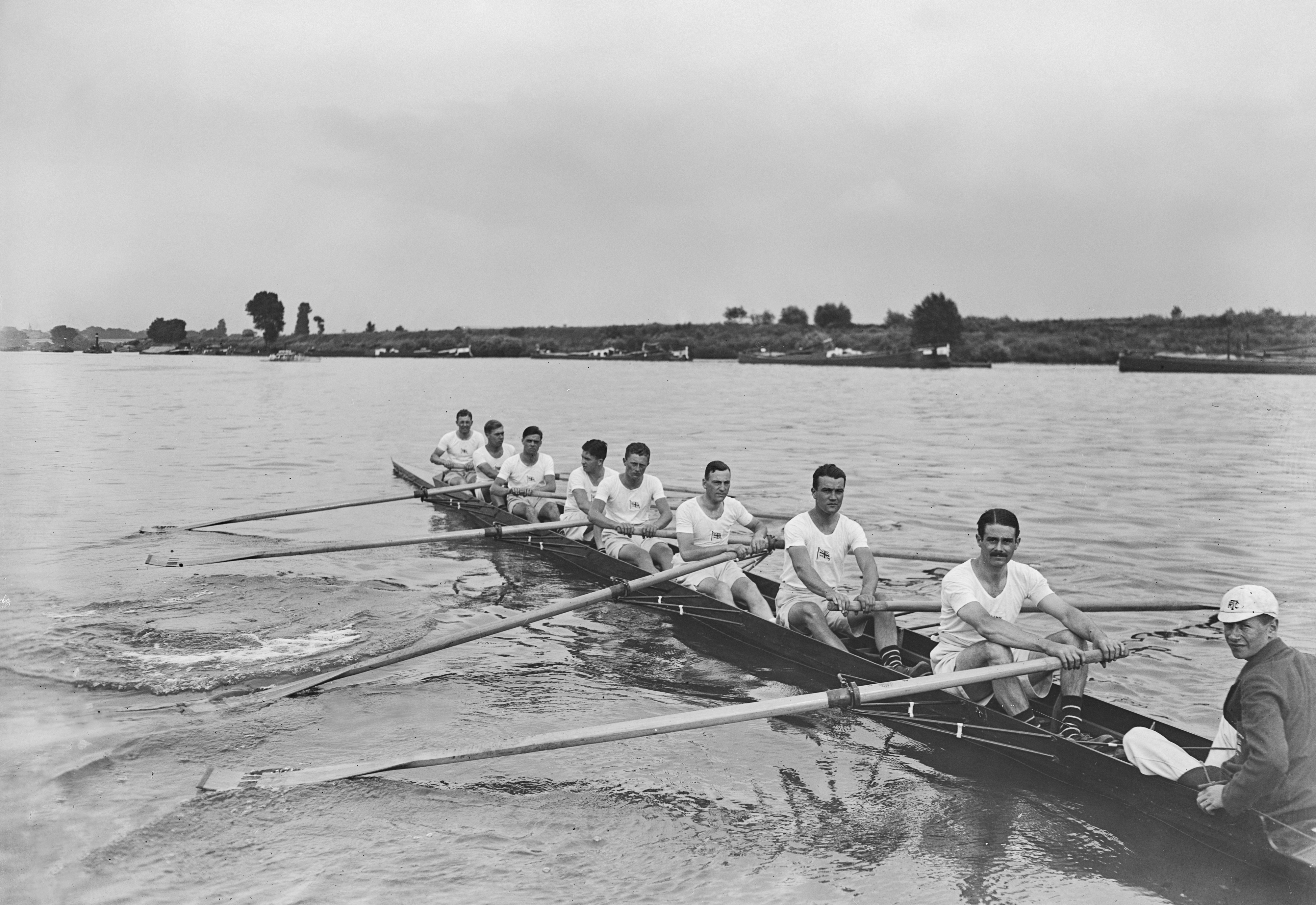 Eight rowers and a coxswain in a team rowing boat on a river during a practice or race. The rowers are all in athletic attire