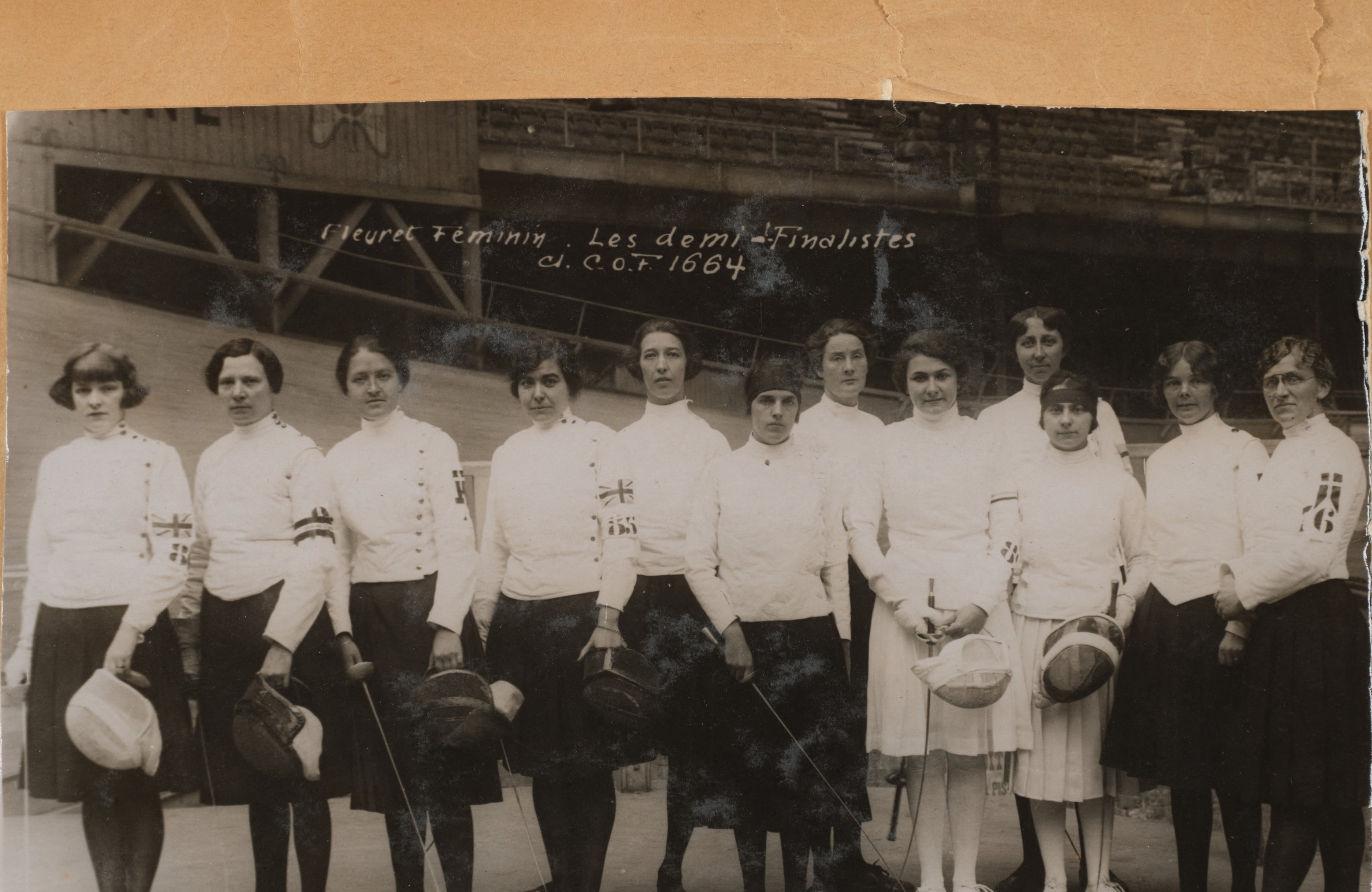 A women&#x27;s historical sports team poses for a group photo, each wearing fencing attire and holding their masks. They stand in front of a wooden structure