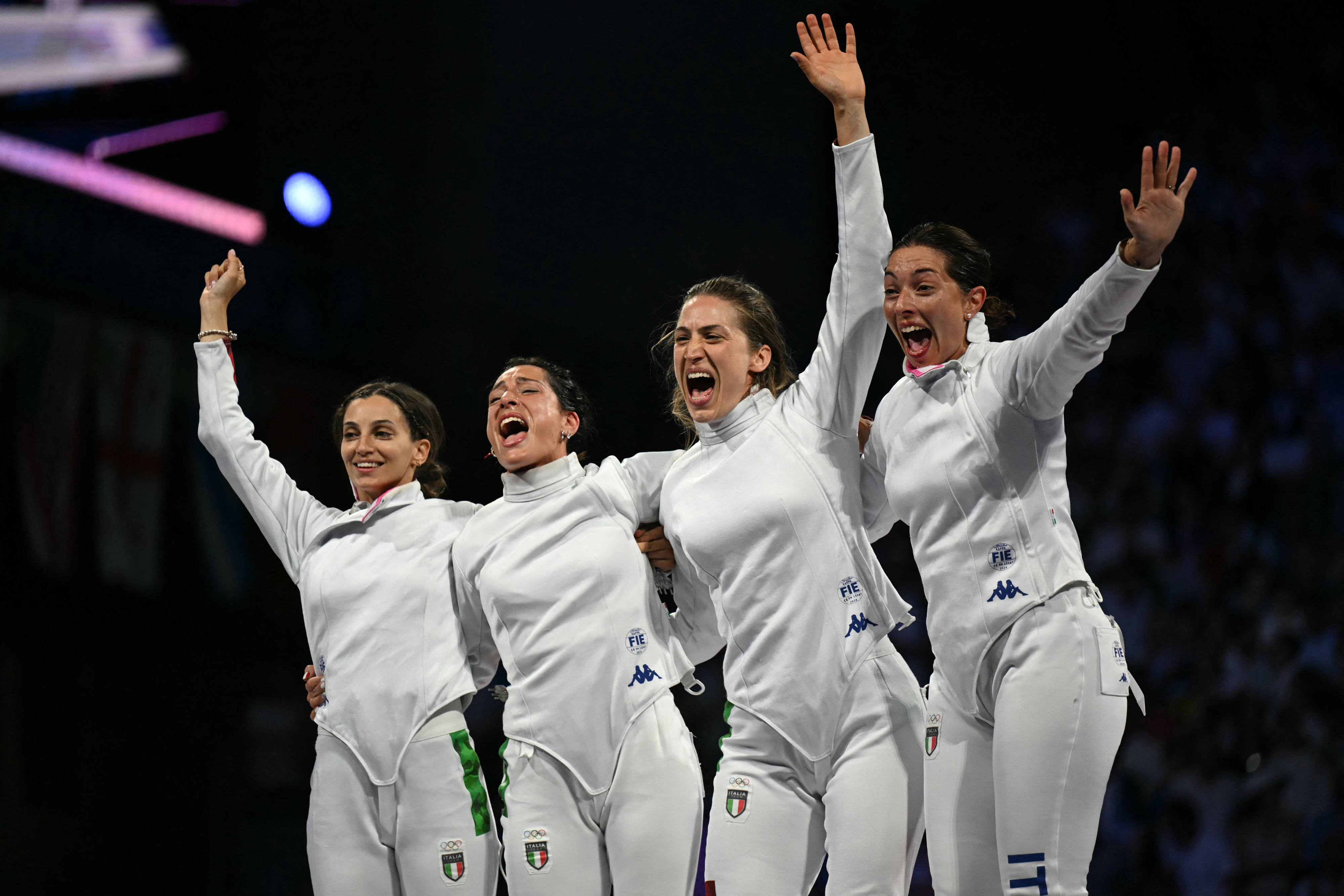 Female fencing team celebrates victory on stage, wearing full fencing uniforms, arms raised in joy