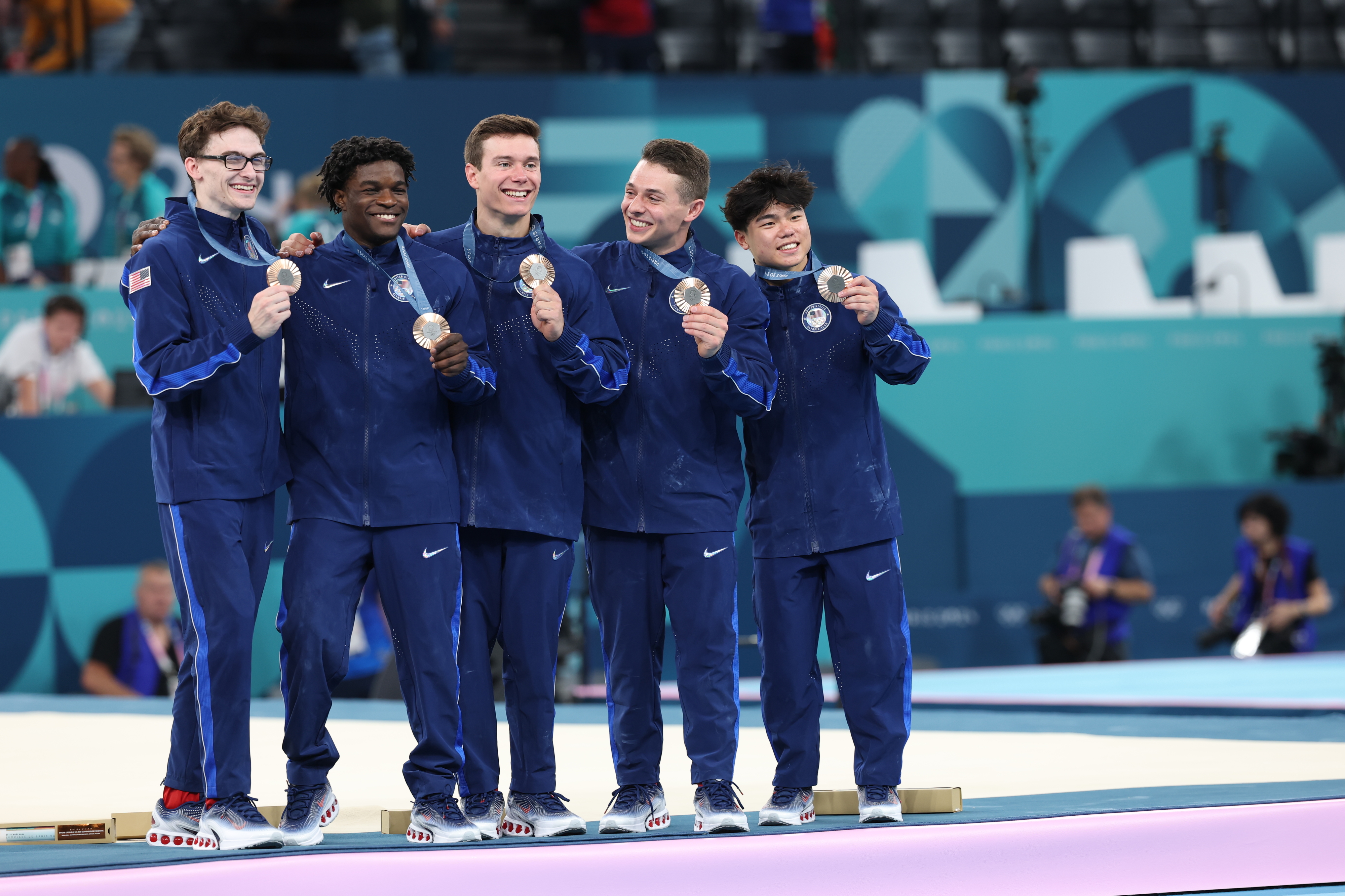 Group of male athletes wearing athletic tracksuits, smiling and holding silver medals, standing on a gymnastic mat at a sports event. Names unknown