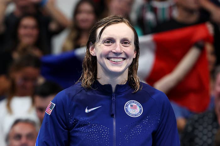 Katie Ledecky, smiling and posing in a sports jacket with the USA flag logo, in front of a blurred crowd with a French flag in the background