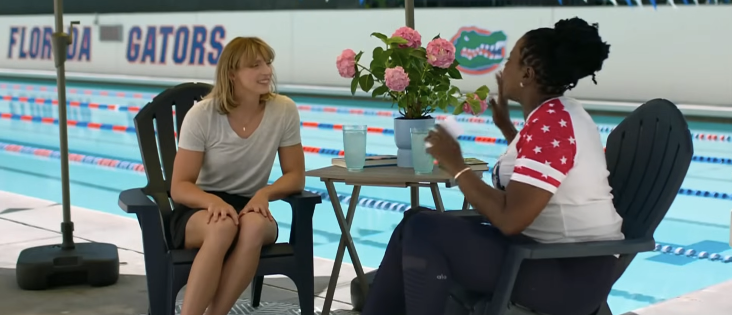 Grace Van Patten and an unidentified person discuss beside an outdoor swimming pool. Grace wears a casual t-shirt and shorts, while the person wears a sporty t-shirt