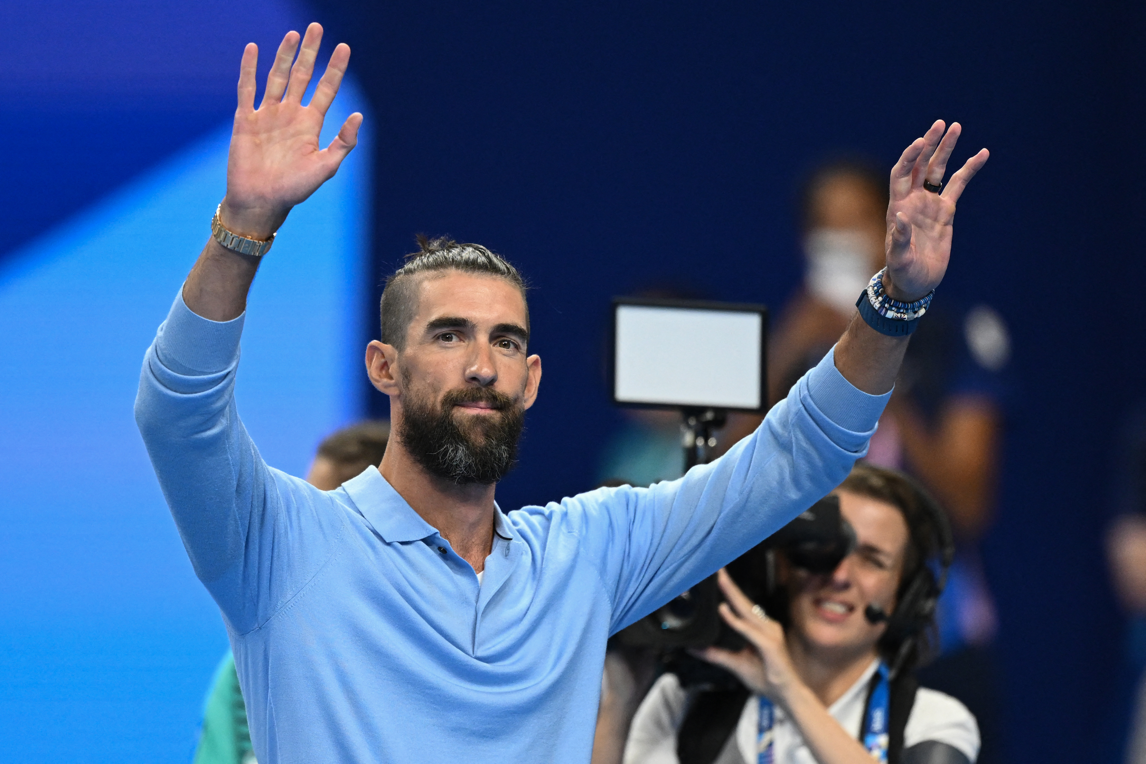 Michael Phelps waves to the crowd wearing a light blue polo and bracelets, with a cameraman and a blurred audience in the background