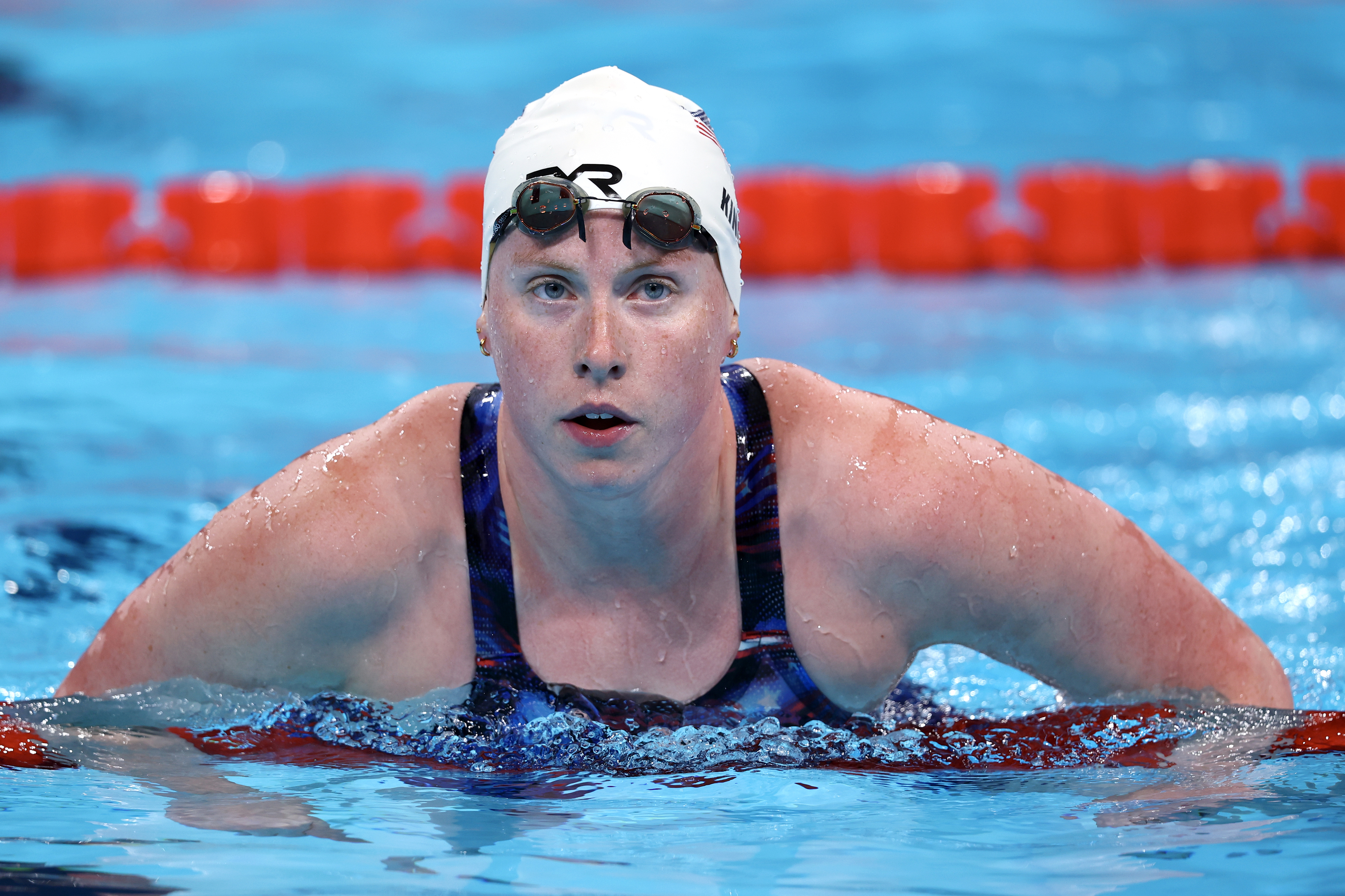 A swimmer, wearing goggles and a swim cap, emerges from a pool during a swim meet