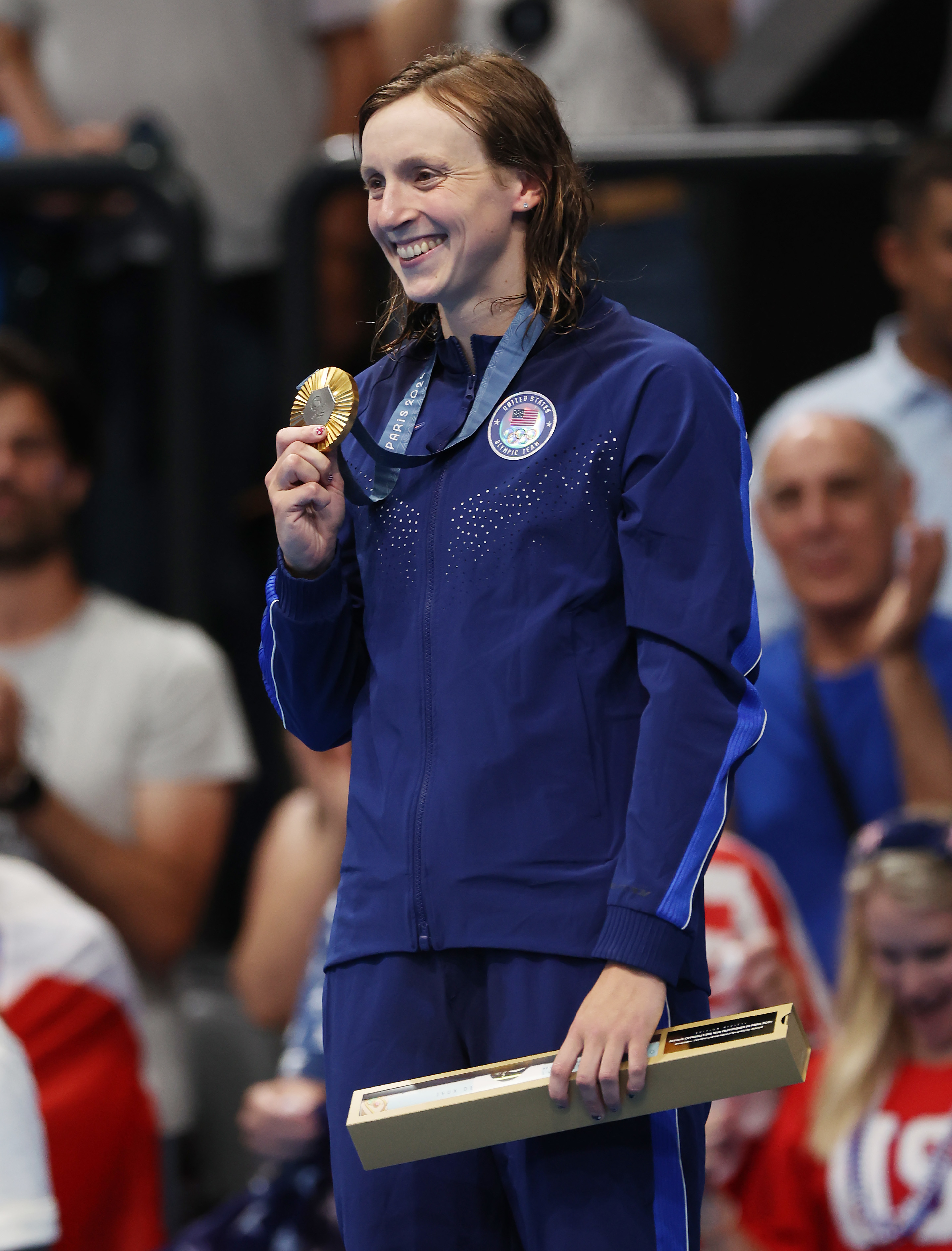 Katie Ledecky, in a tracksuit, smiles and holds an award medal with one hand and a boxed certificate with the other during a ceremony in a sports arena