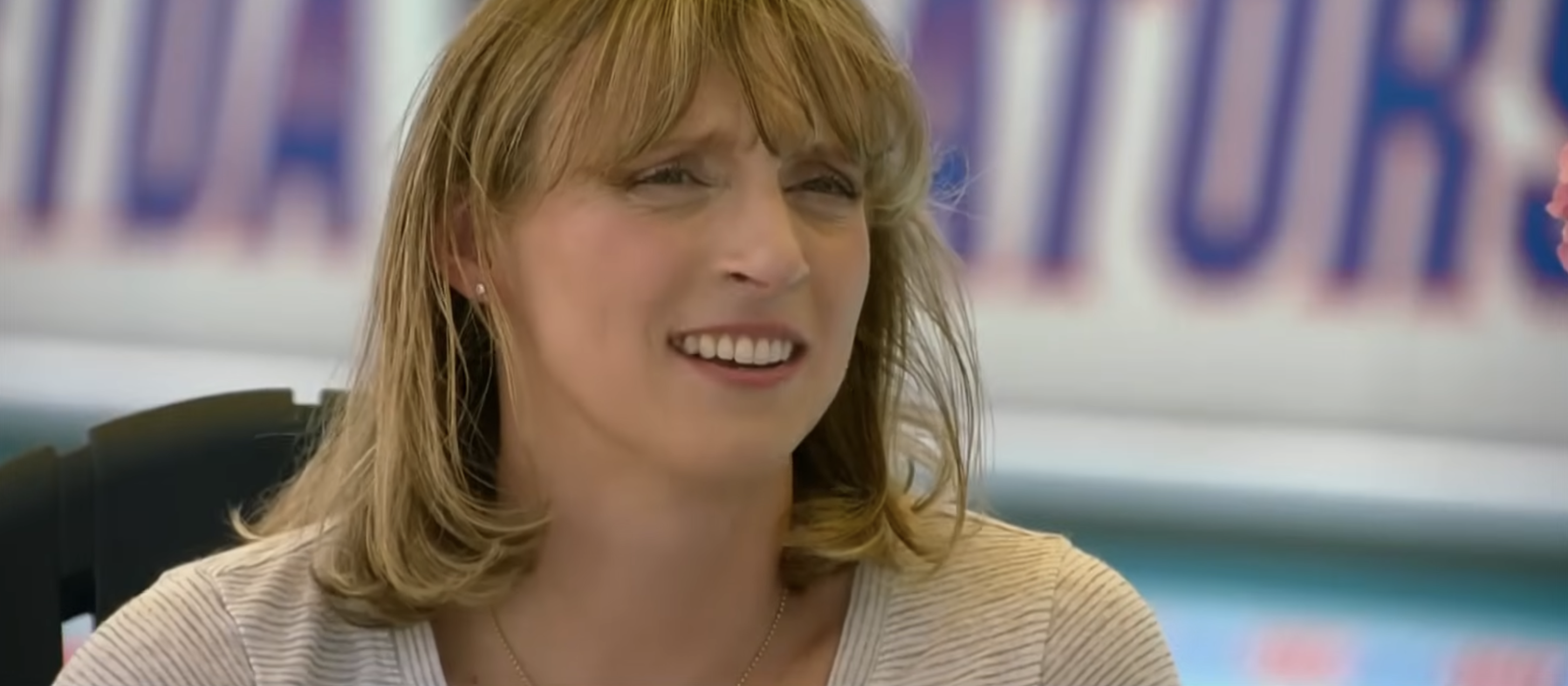 A woman with shoulder-length hair is smiling slightly. She is wearing a striped top and a necklace. The background is blurred with indistinct text