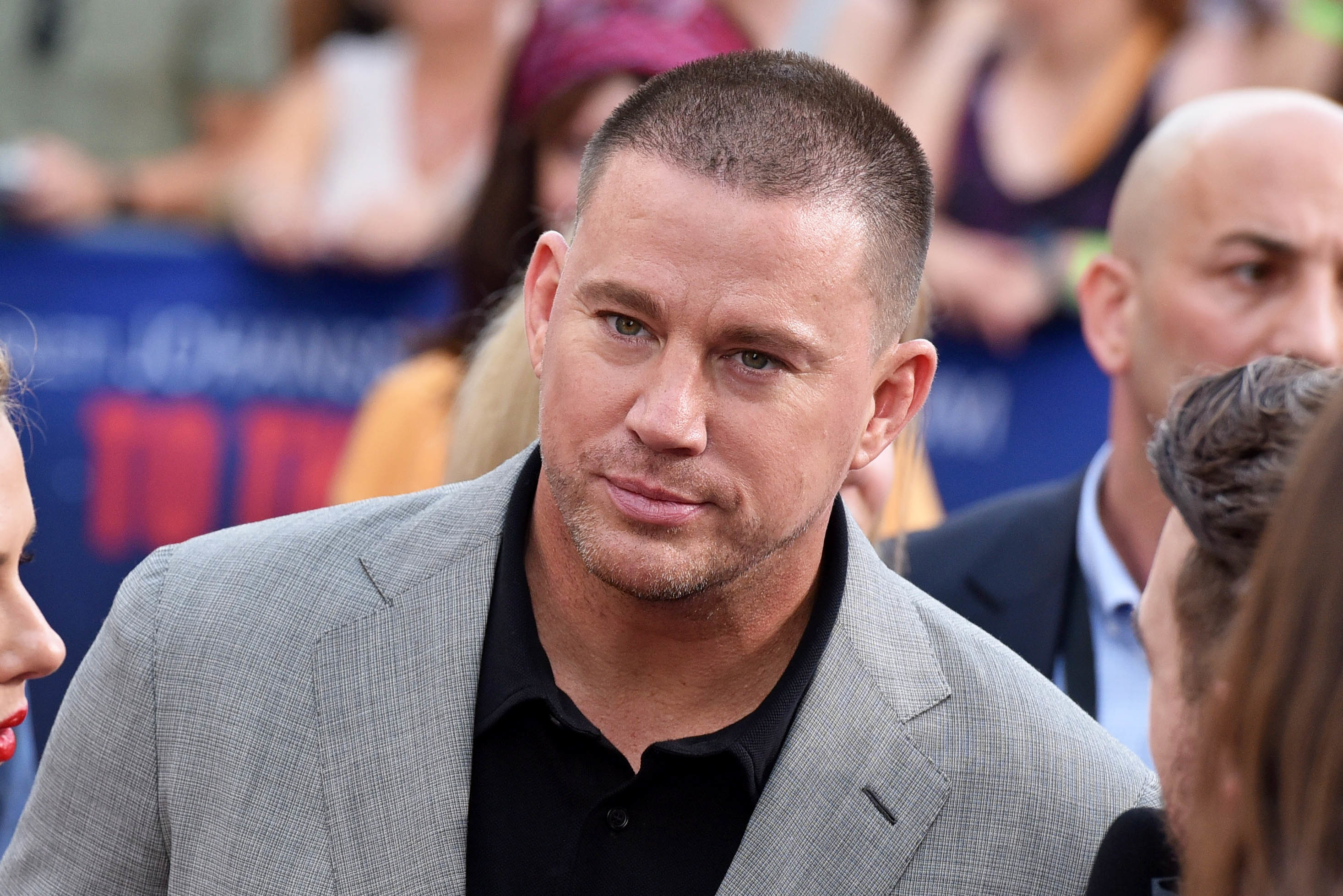 Channing Tatum is wearing a grey suit and black shirt while on a red carpet event. People are visible in the background