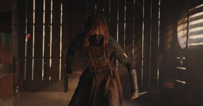 Person in an action pose, dressed in armor with a dark mask, in a dimly lit wooden building interior