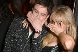 Barry Keoghan and Olivia Rodrigo pose on the red carpet, playfully covering their mouths with their hands. Rodrigo wears a shiny black dress with sheer elements