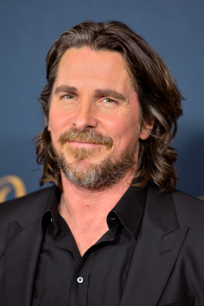 Christian Bale, with medium-length hair and a beard, wearing a suit and dress shirt, smiles at a formal event