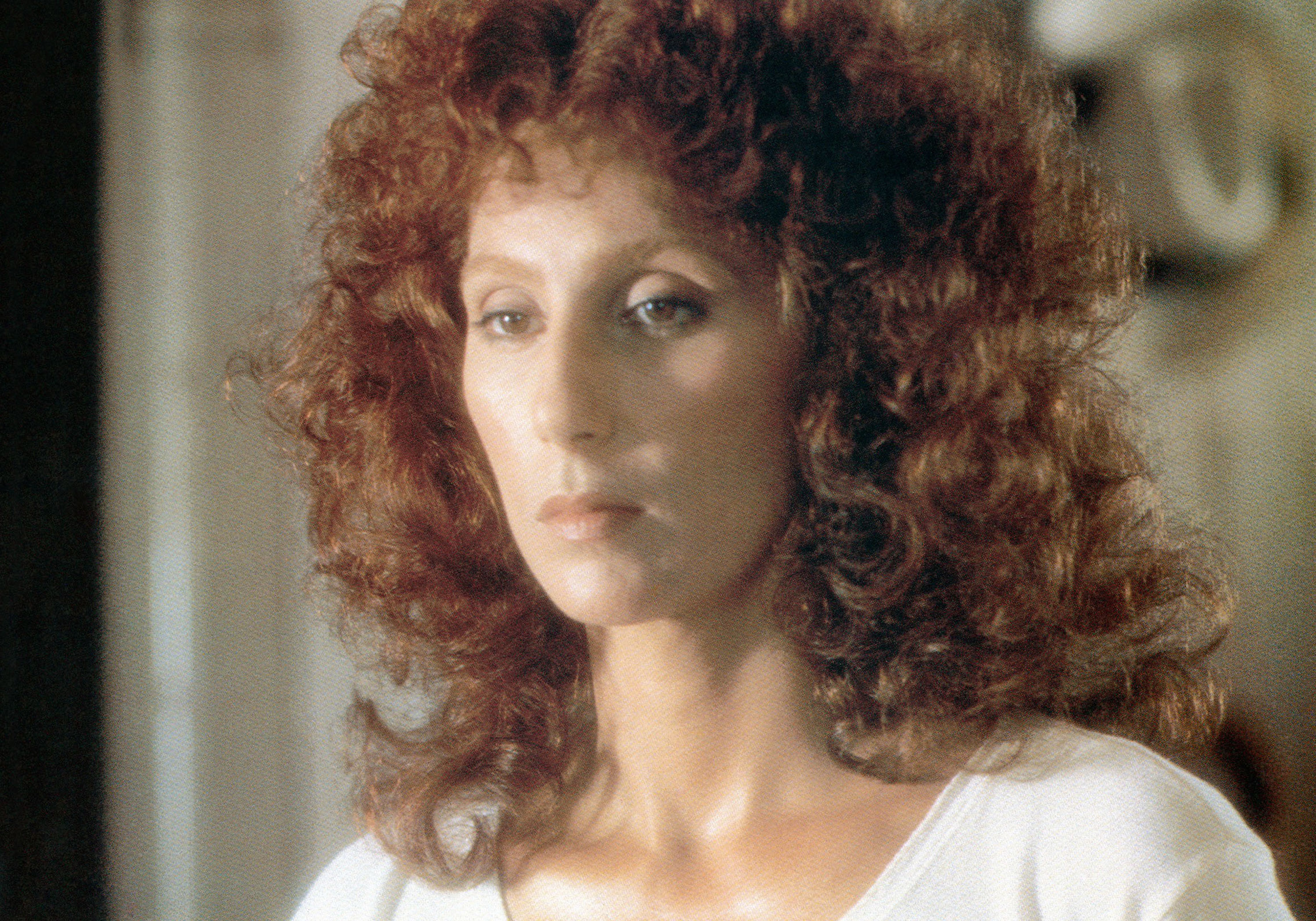 Cher with voluminous curly hair wearing a simple light-colored top looks serious and contemplative