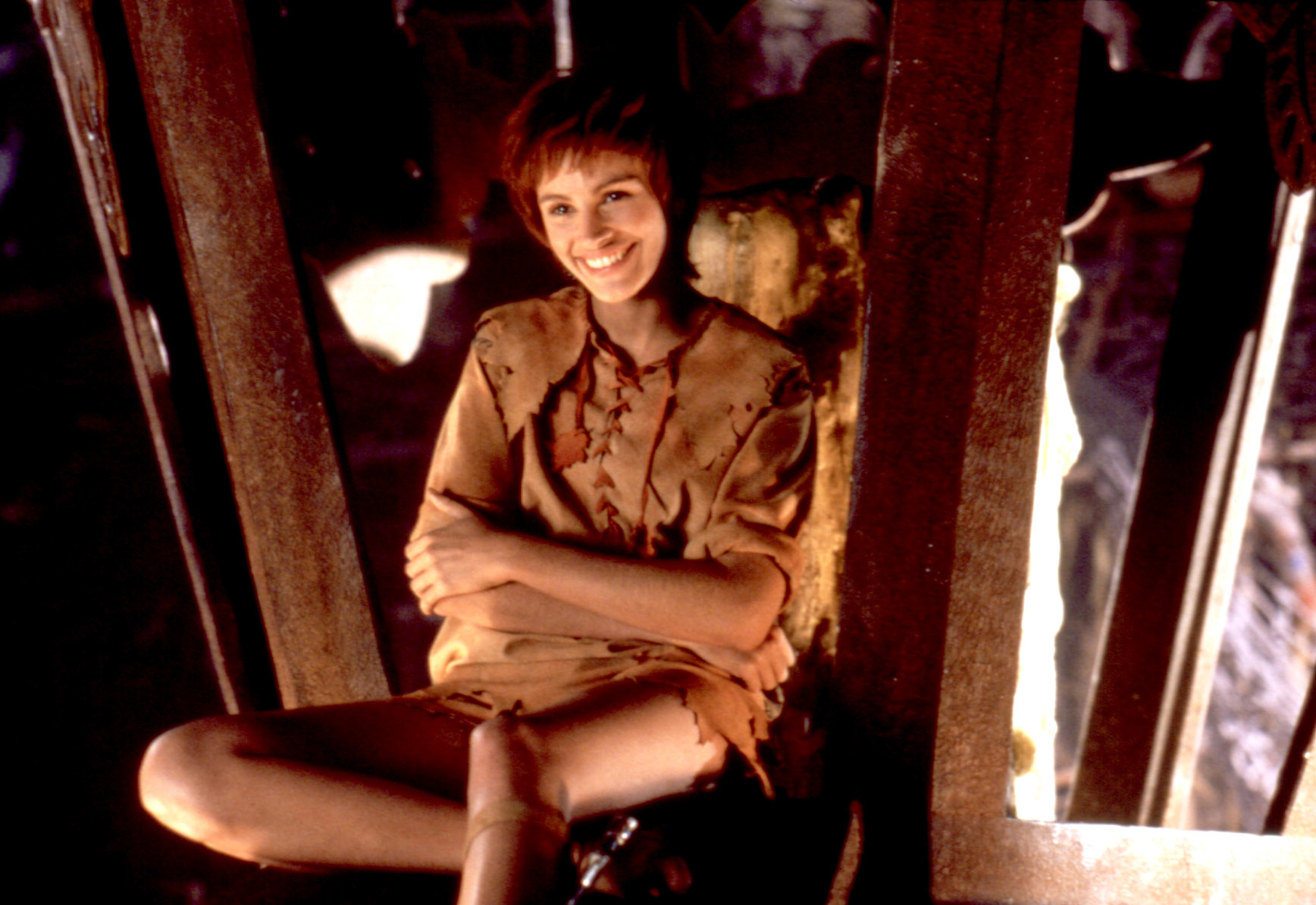 Julia Roberts as Tinkerbell dressed in rustic, medieval-style clothing, sitting with legs crossed, smiling in a film scene