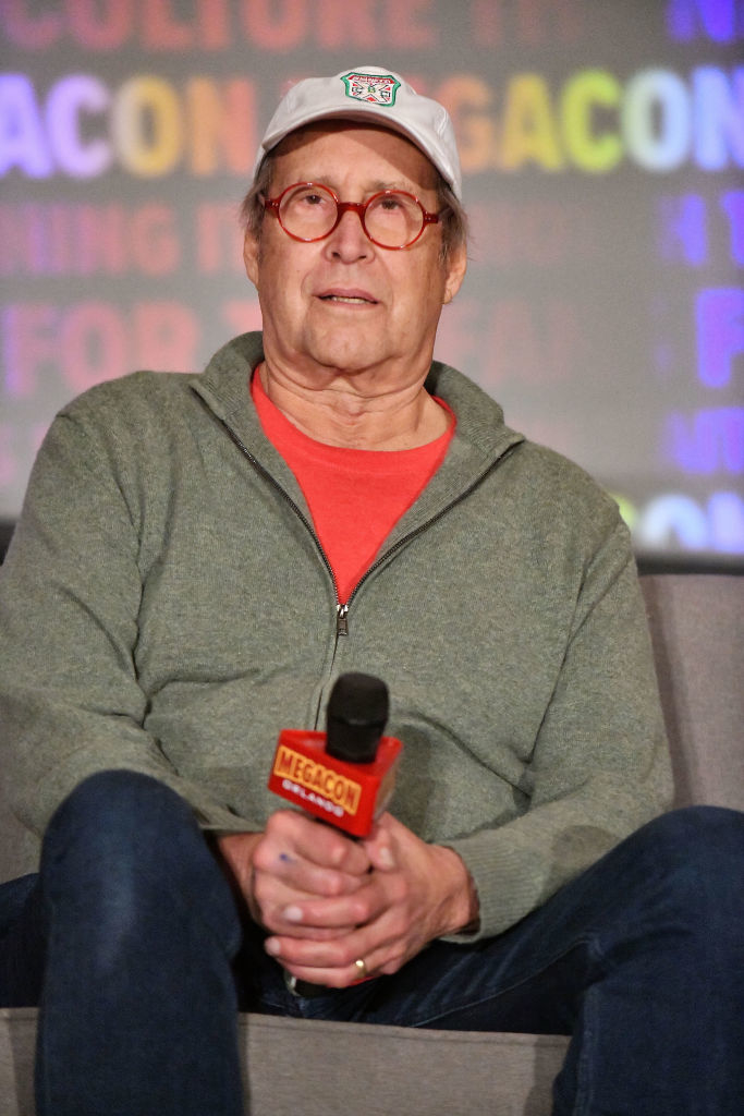 Chevy Chase sits on a couch at an event, holding a microphone with a &quot;MegaCon&quot; logo. He is wearing a casual outfit with a cap