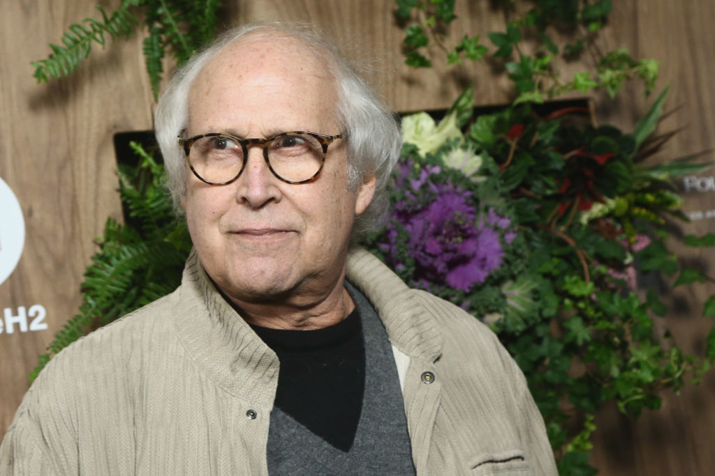 Chevy Chase wearing glasses and a casual outfit, standing in front of a floral and greenery backdrop