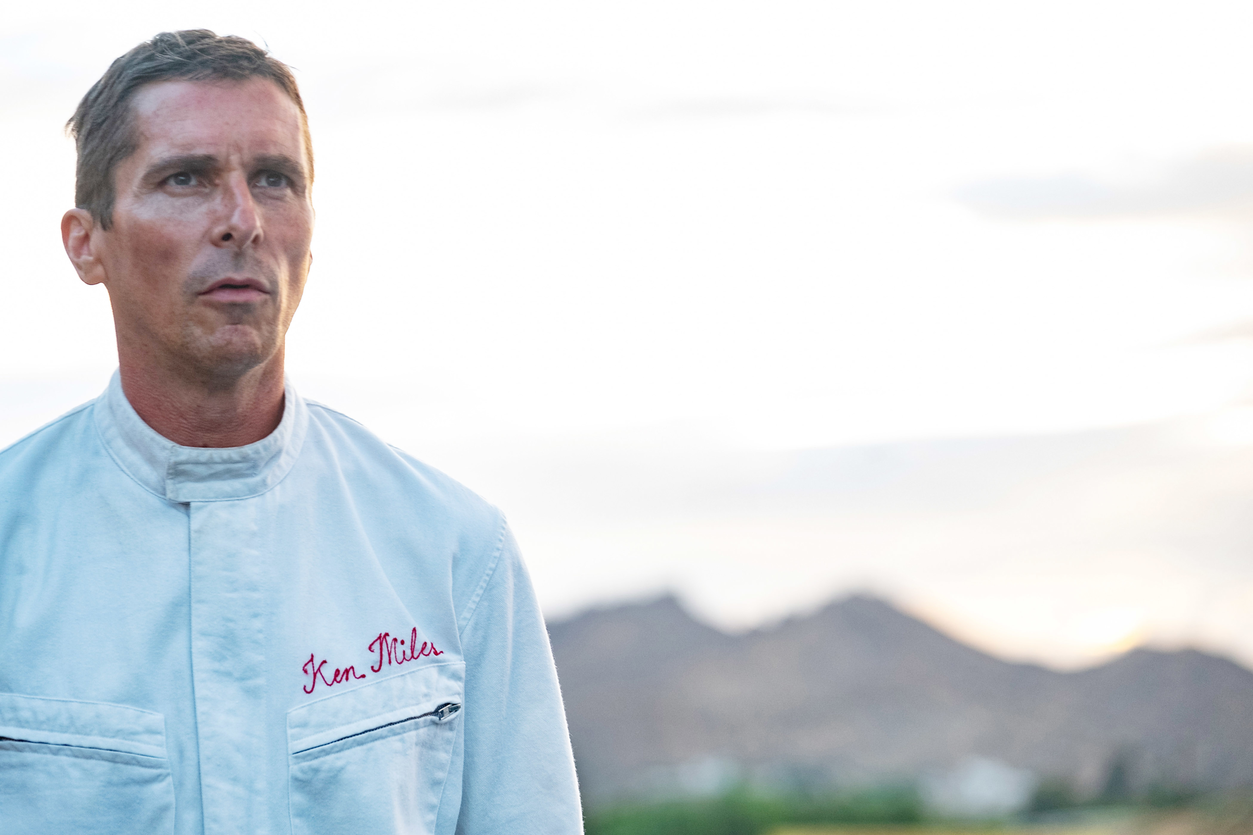 Christian Bale stands outdoors in a jacket embroidered with &quot;Ken Miles,&quot; with blurred mountains in the background