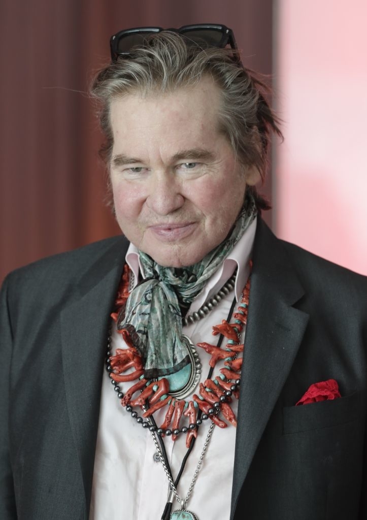 Val Kilmer wearing a suit with a large beaded necklace and scarf, and glasses on his head, smiling at an event