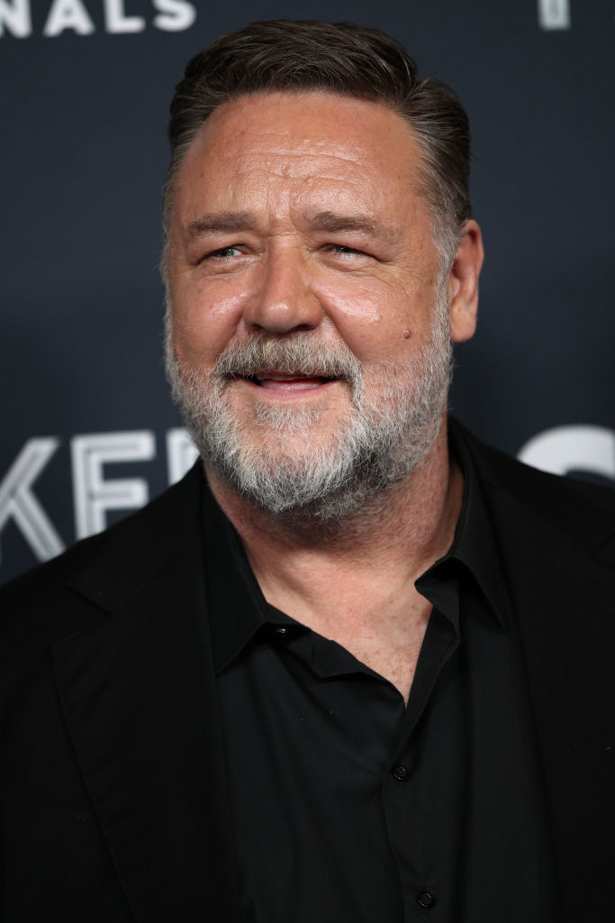 Russell Crowe smiles on the red carpet, dressed in a dark shirt and matching blazer