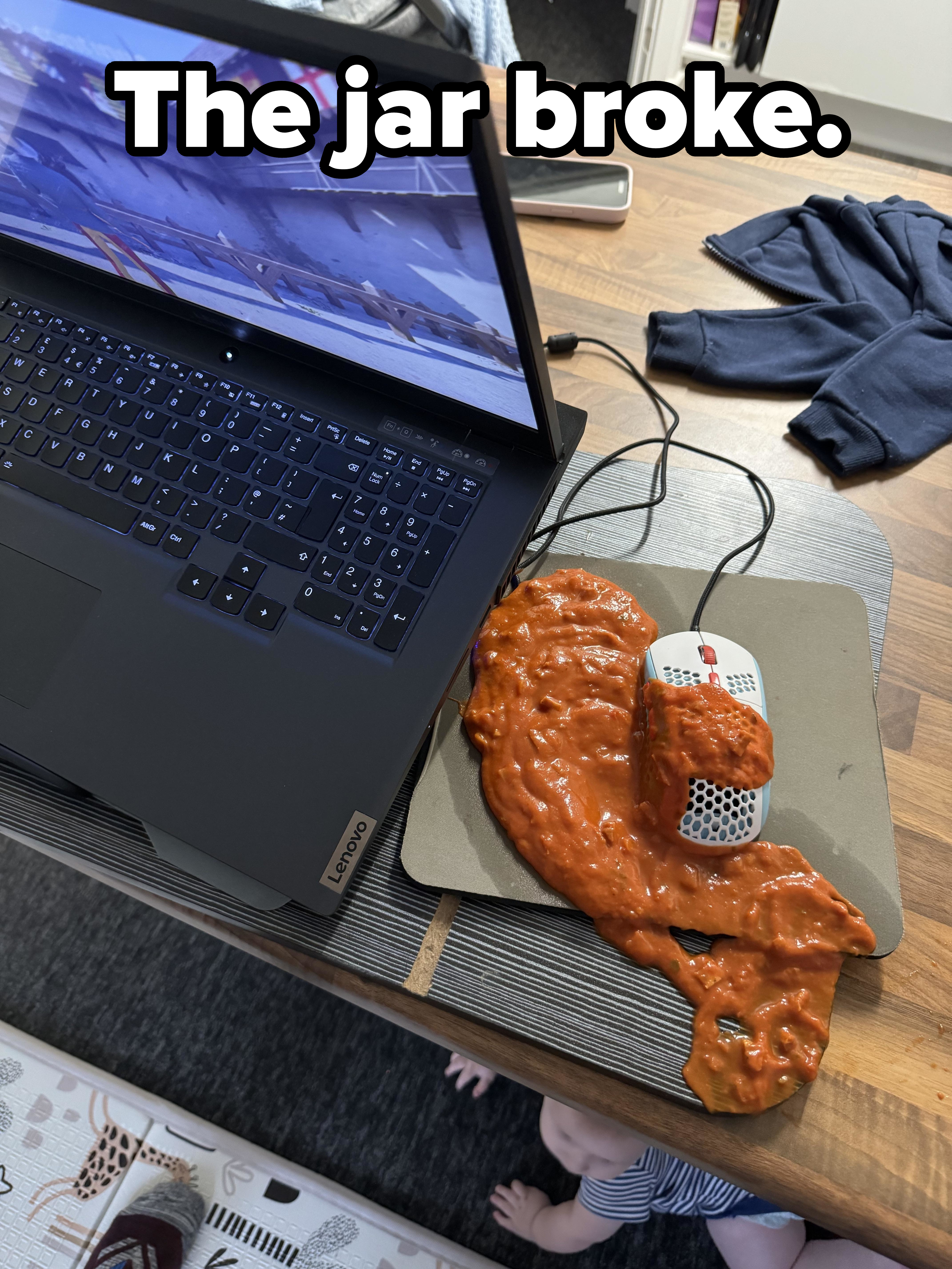 A laptop on a table with a mouse covered in what appears to be pasta sauce next to it. A baby is seen crawling underneath the table