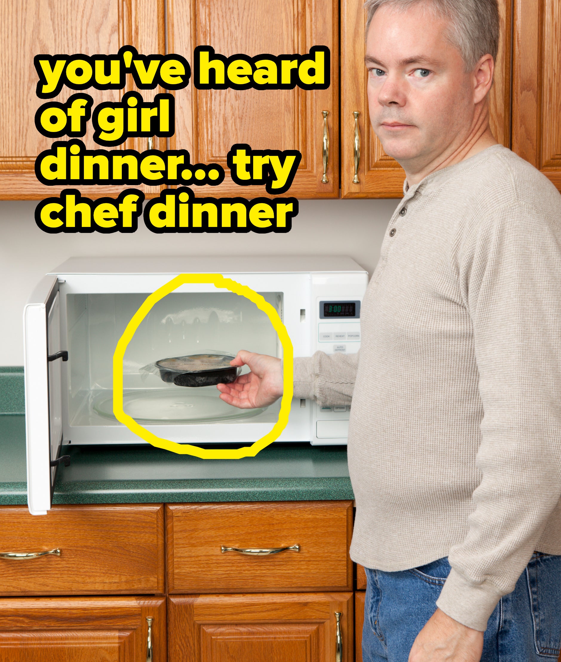 A man with gray hair and a light sweater holds a pan while using a microwave in a kitchen with wooden cabinets