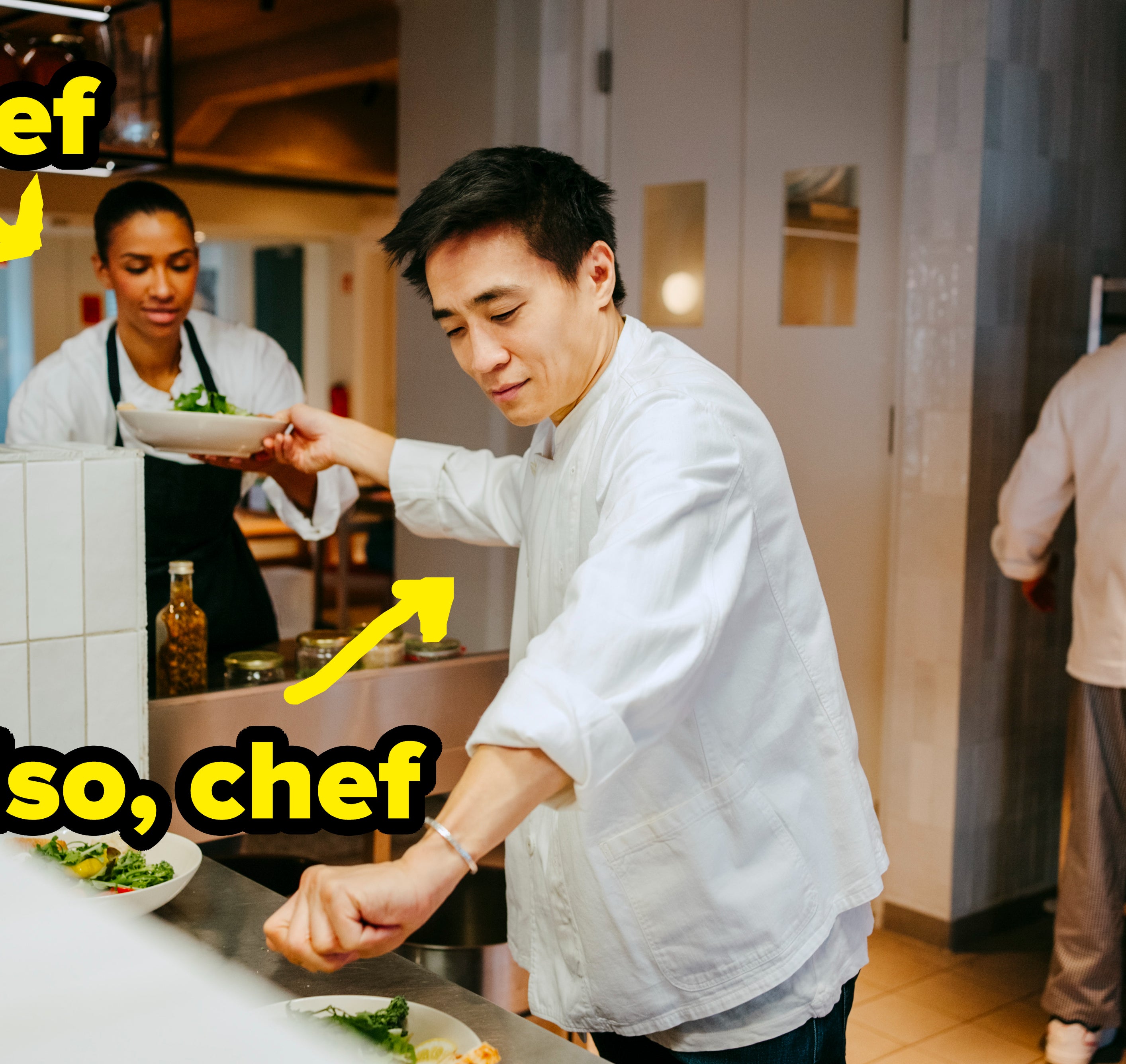 Chefs in a modern kitchen, preparing dishes on plates. A male chef is placing greens on a plate while a female chef places a dish on the counter. Another chef is in the background