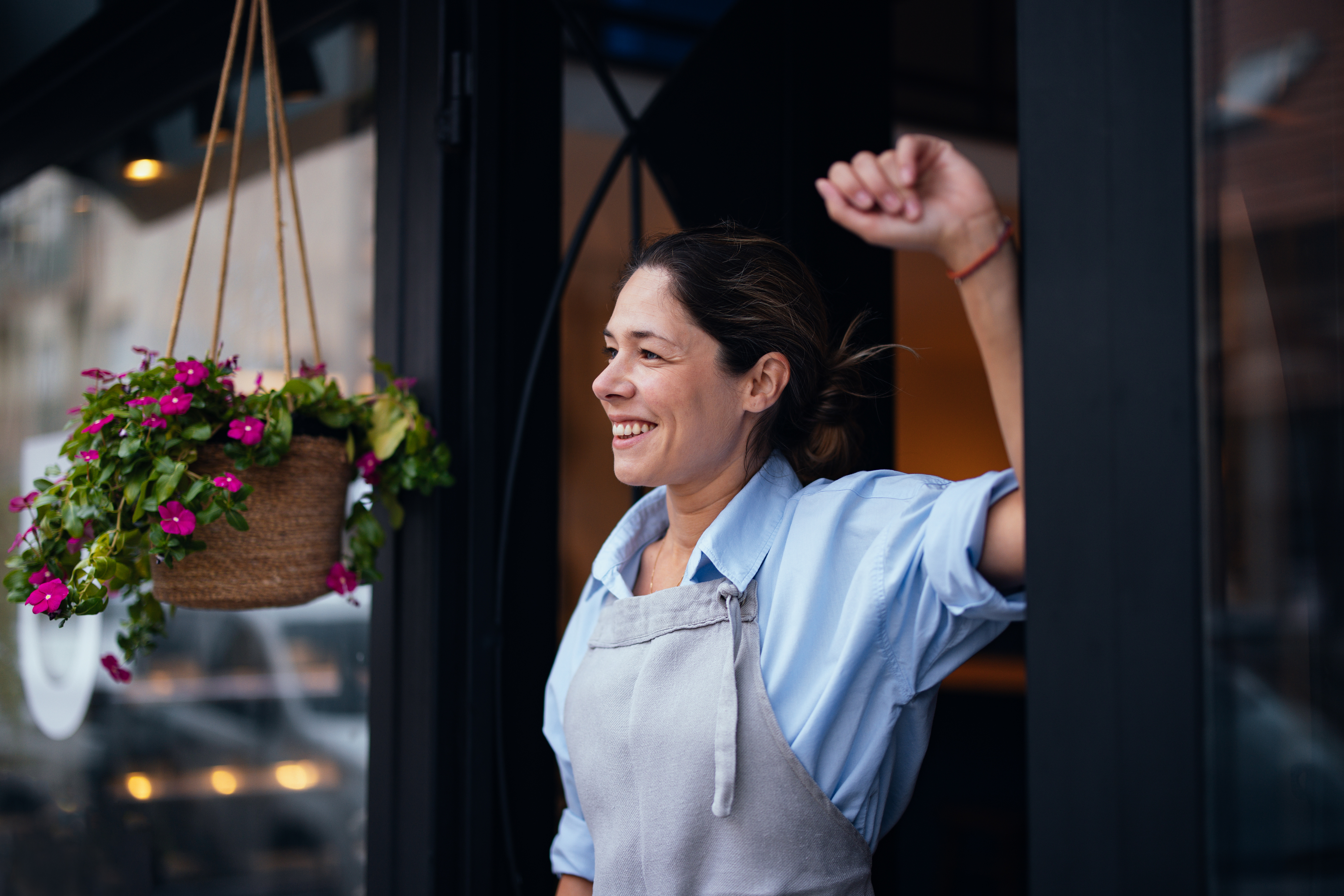 A woman wearing a light blue shirt and apron smiles while standing at the entrance of a bakery, with a hanging flower basket nearby