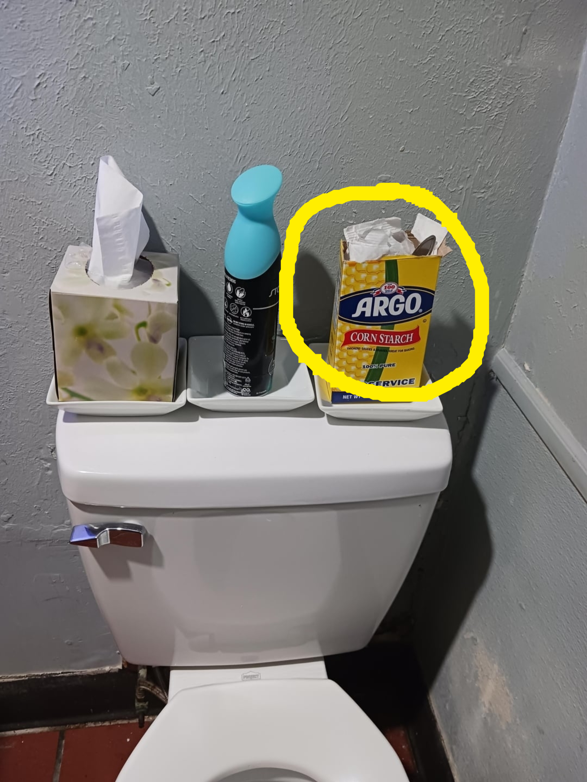 Toilet with a tissue box, air freshener, and a box of Argo corn starch on the tank lid