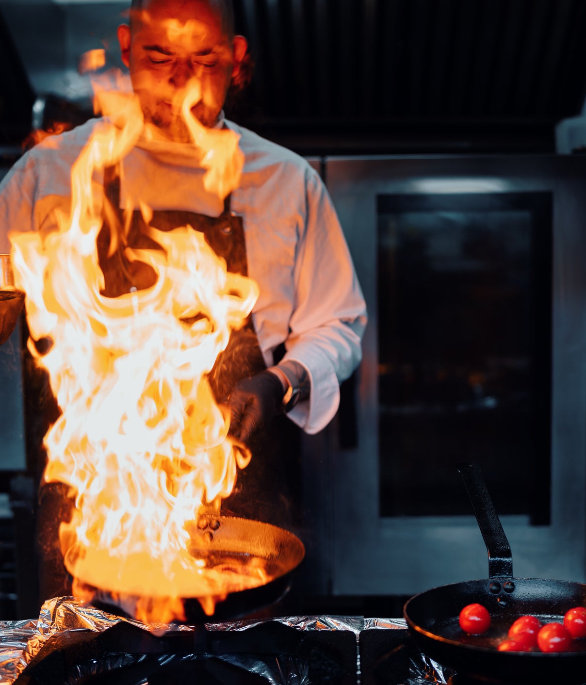 A chef in a professional kitchen uses a burner to create a large flame over a skillet. Another pan with tomatoes is visible next to him