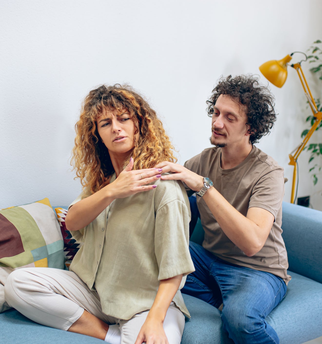 A woman and a man sit on a couch; the man appears to massage the woman&#x27;s shoulder as she looks at her back with discomfort