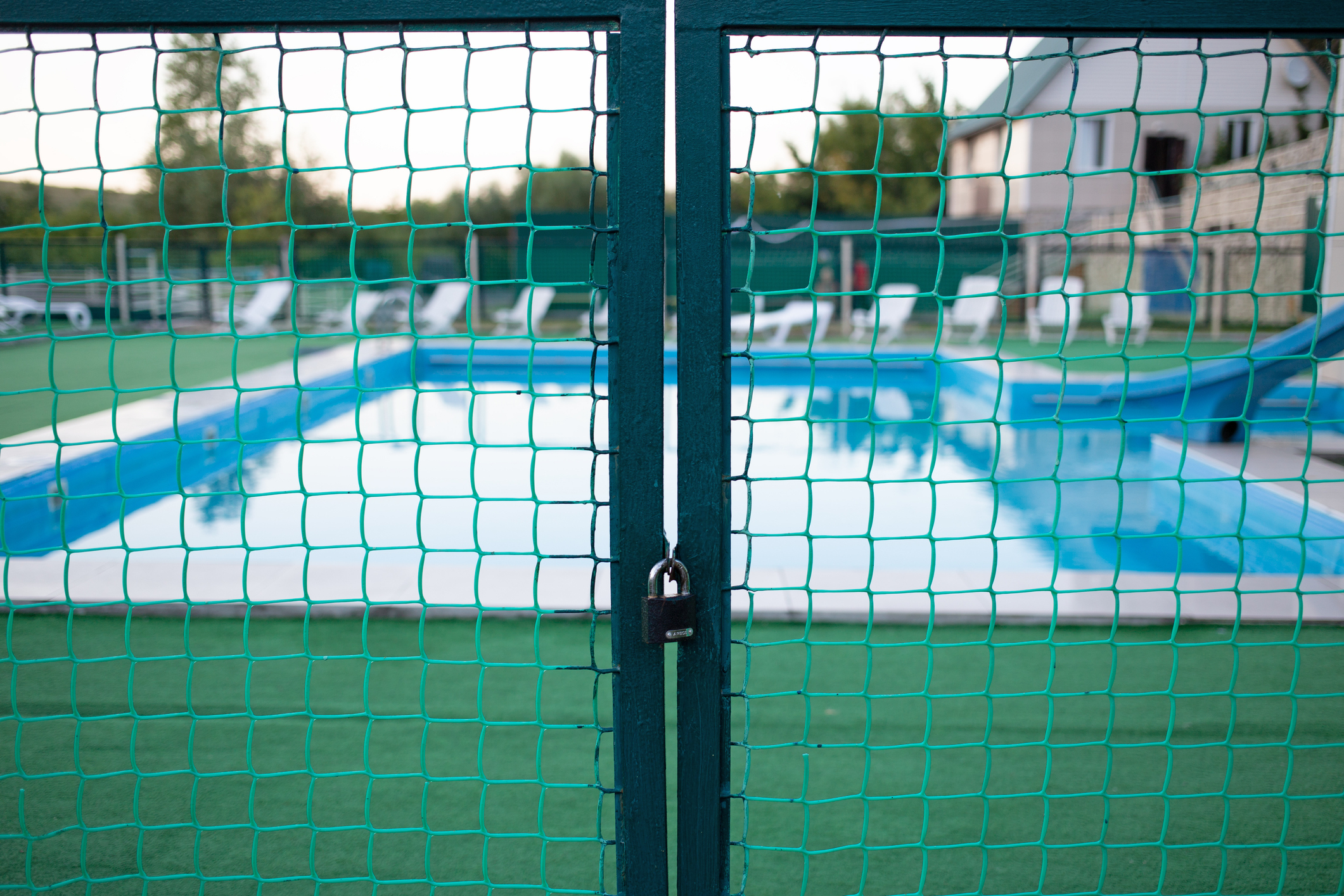 A locked gate with green netting blocks access to an empty swimming pool area with lounge chairs and a building in the background