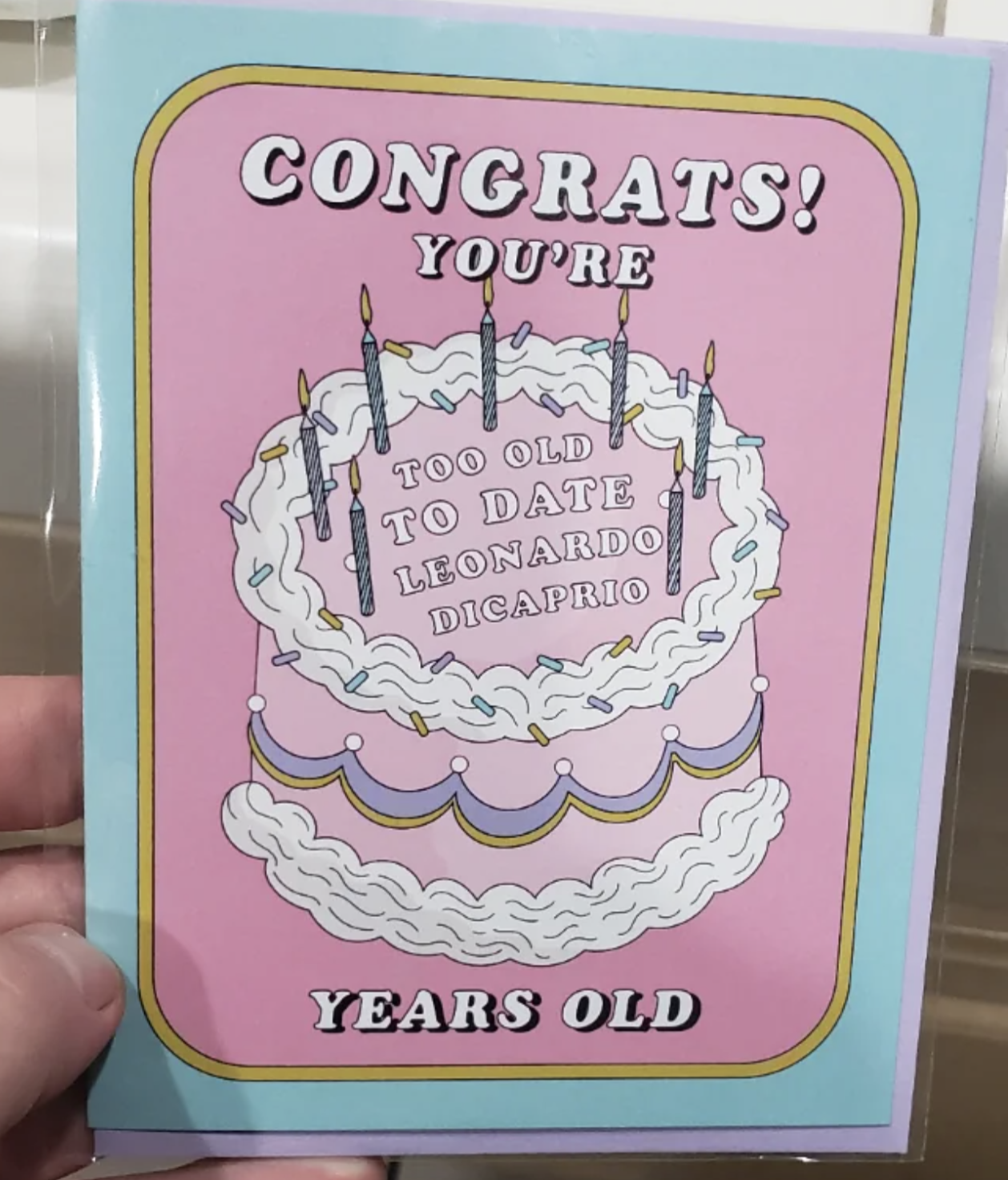 A hand holds a greeting card with a drawing of a birthday cake with candles. The card reads: &quot;CONGRATS! YOU&#x27;RE TOO OLD TO DATE LEONARDO DICAPRIO YEARS OLD.&quot;