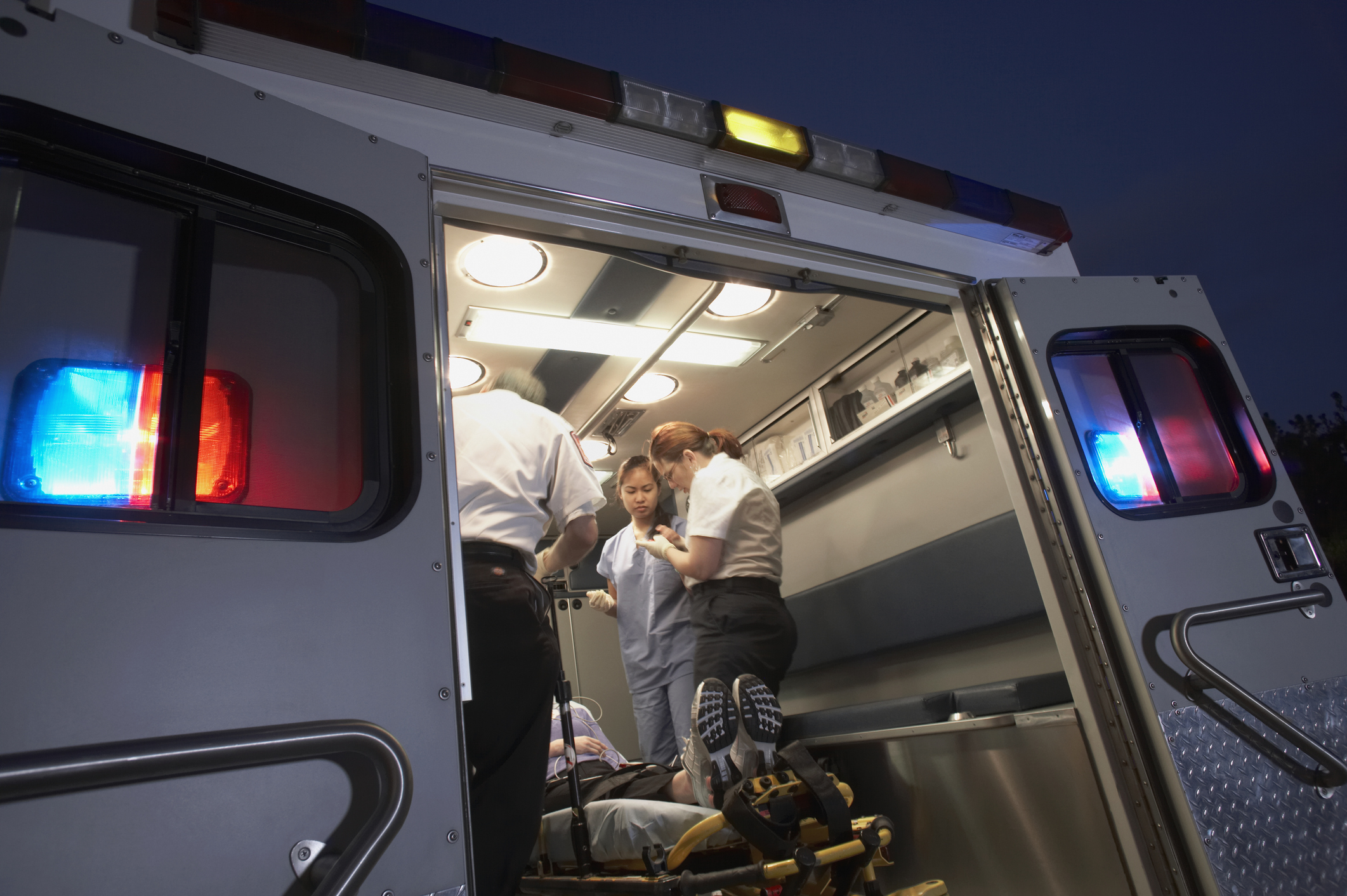 Paramedics and a doctor attend to a patient on a stretcher inside an ambulance. Red and blue lights are visible on the ambulance