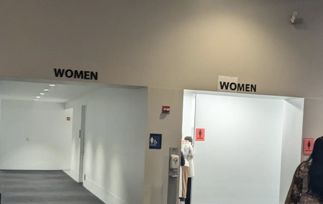 Two restroom entrances labeled &quot;Women&quot; with one having a hand sanitizer station nearby; a person partially visible on the right