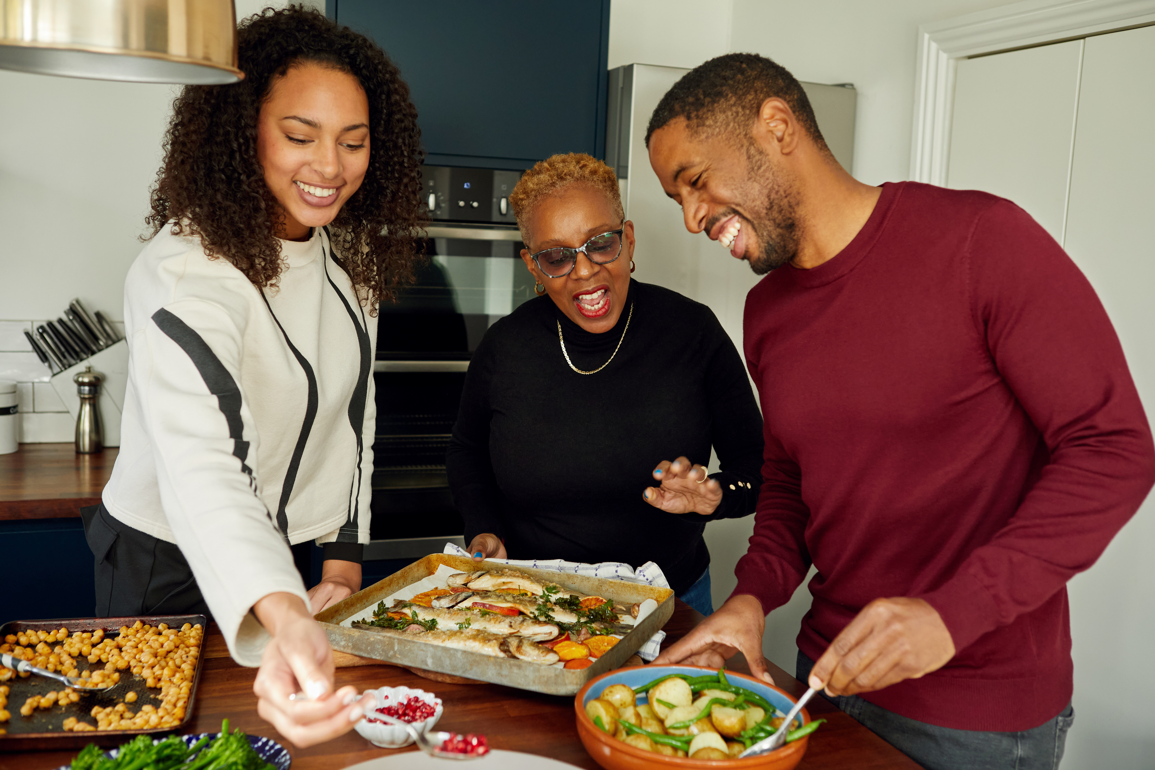 Three adults, all smiling and engaged in preparing food together in a modern kitchen. They appear to be enjoying making a meal