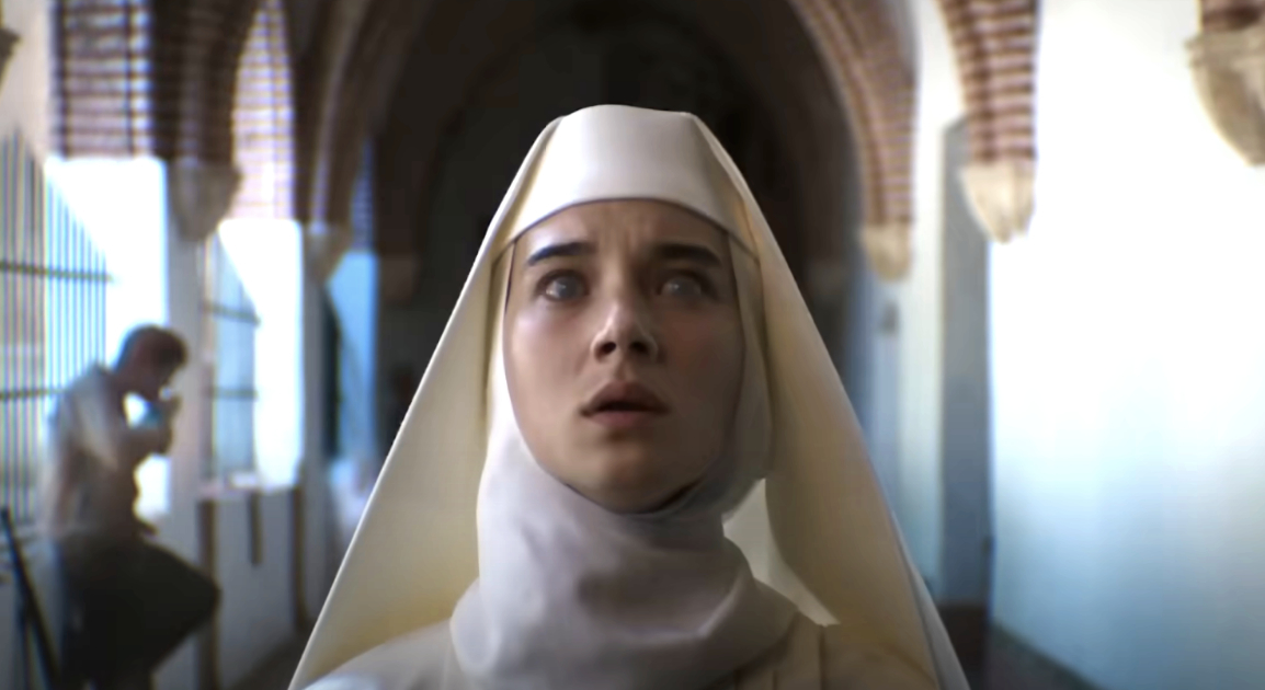 A woman dressed as a nun with a white habit looks upward with a concerned expression in an arched hallway. Another person is seated in the background