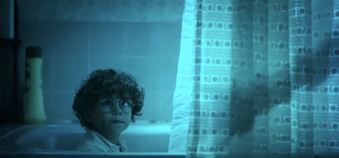 Young boy with curly hair and glasses inside a bathtub, looking towards a patterned shower curtain as an arm reaches for him