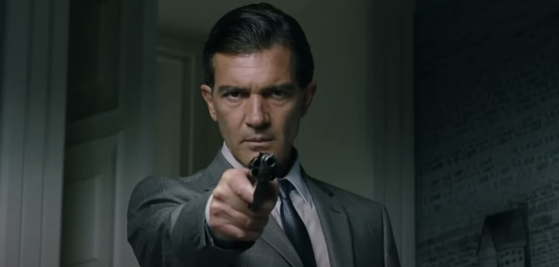 Antonio Banderas points a gun in a tense scene, dressed in a suit and tie