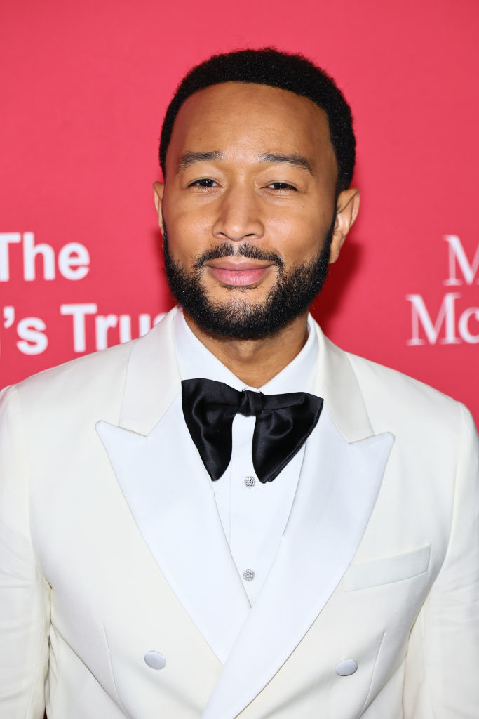 John Legend in a white tuxedo with a black bow tie, at an event with a red backdrop