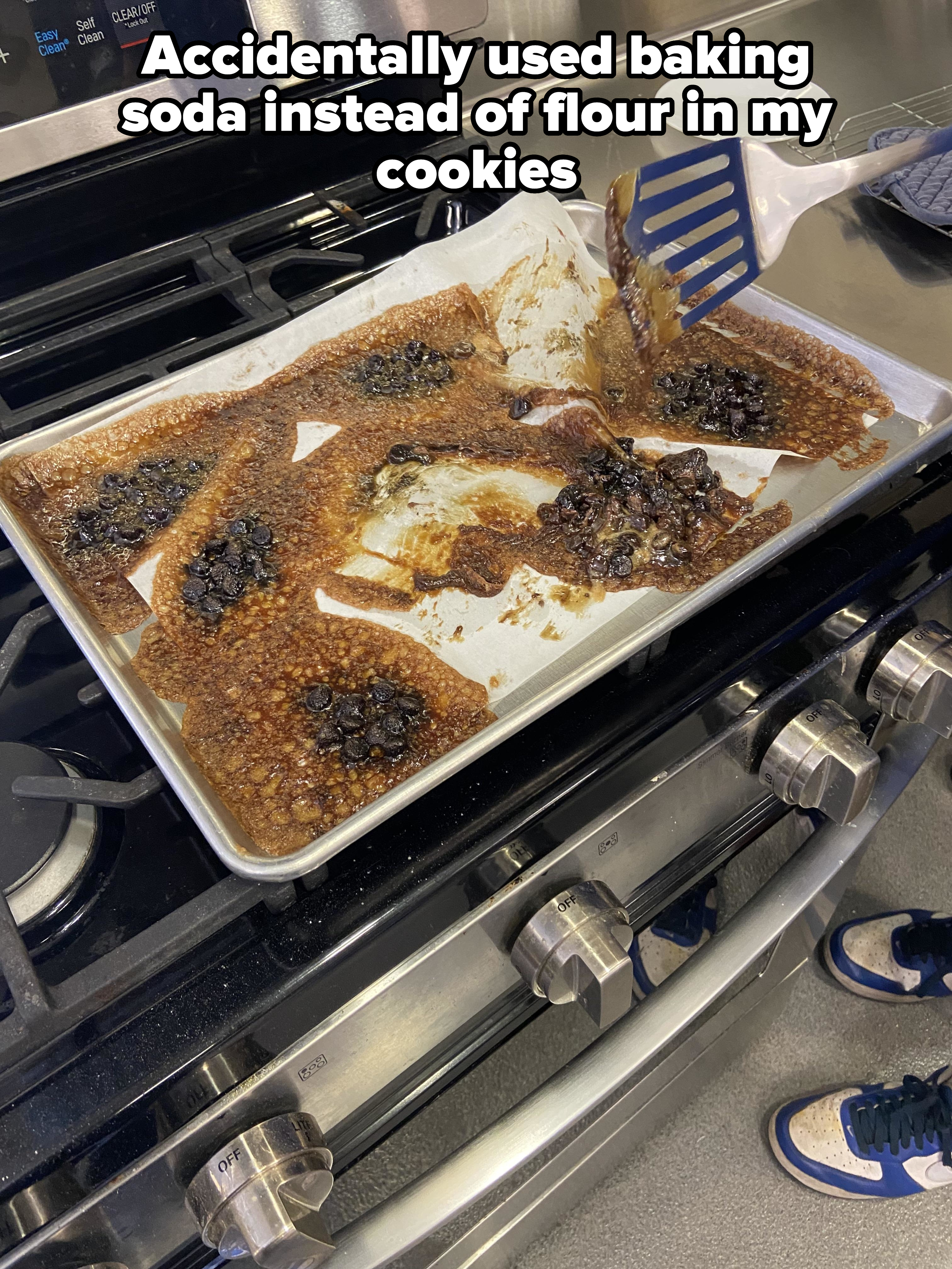 Burnt, collapsed cookies on a baking sheet being scooped up with a spatula over a gas stove, some cookies are partially intact, and partially charred