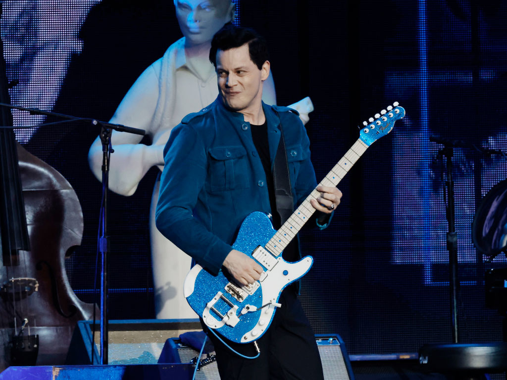 Jack White performs on stage, playing a blue electric guitar, with a statue in the background