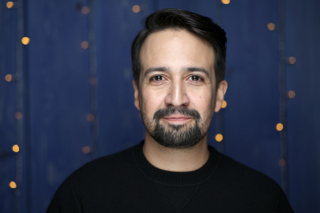 Lin-Manuel Miranda is smiling, wearing a dark sweater, with a backdrop of blue and twinkling lights