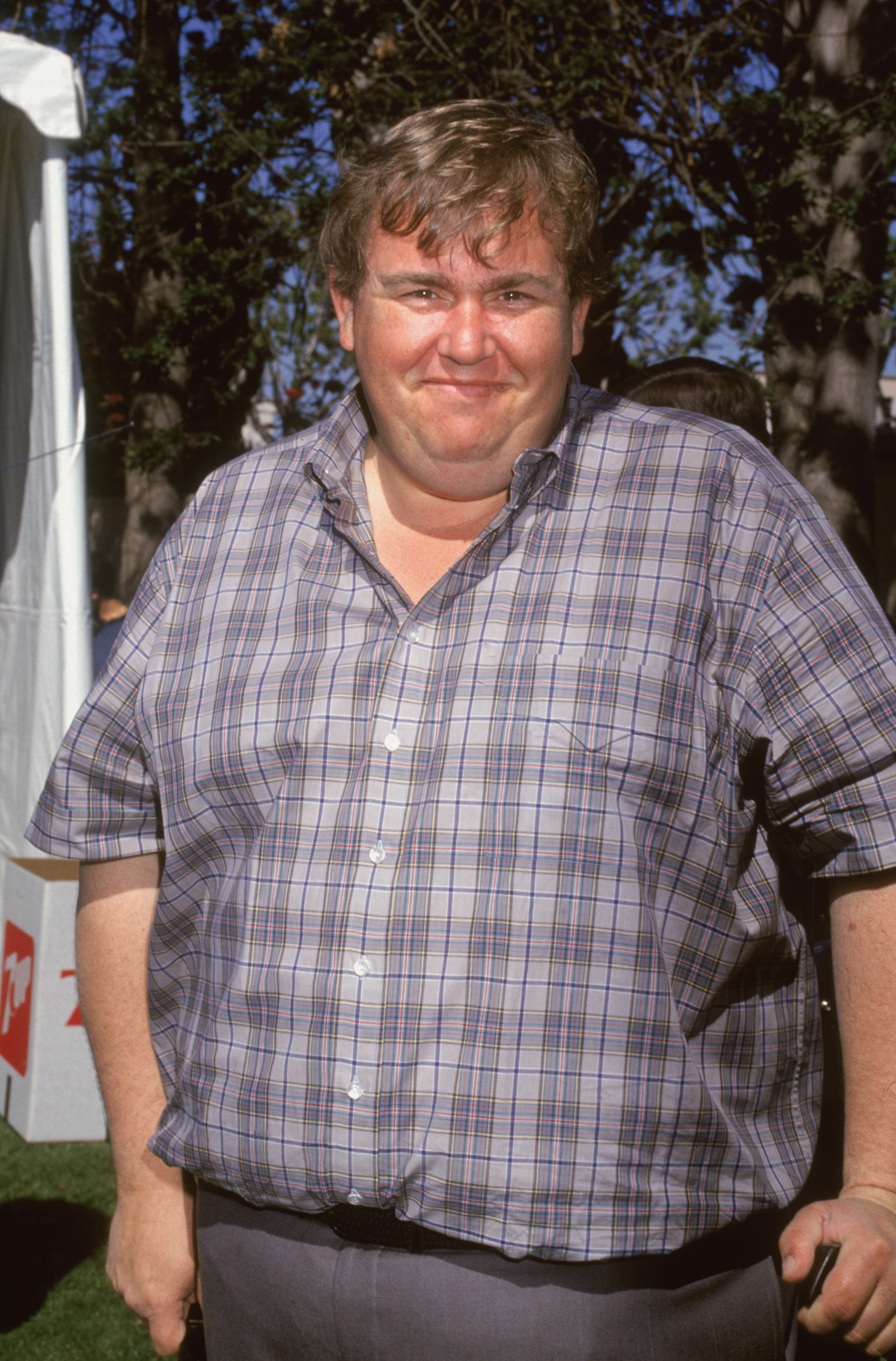 John Candy, wearing a checked shirt and dark pants, stands outdoors at an event, smiling at the camera. Trees and a tent are visible in the background